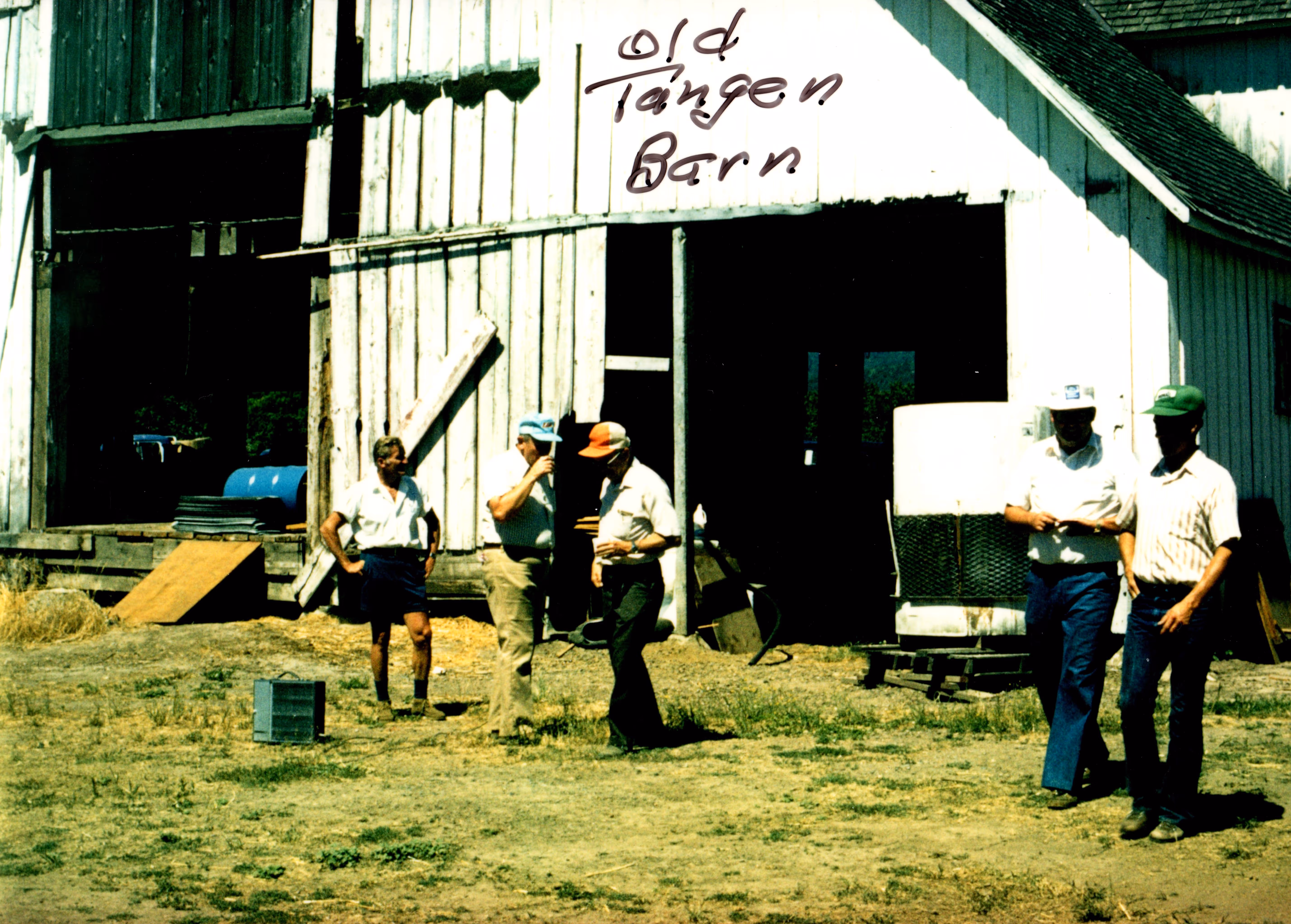 Five men standing and talking in front of a white wooden barn with 'Old Tangen Barn' handwritten on it.