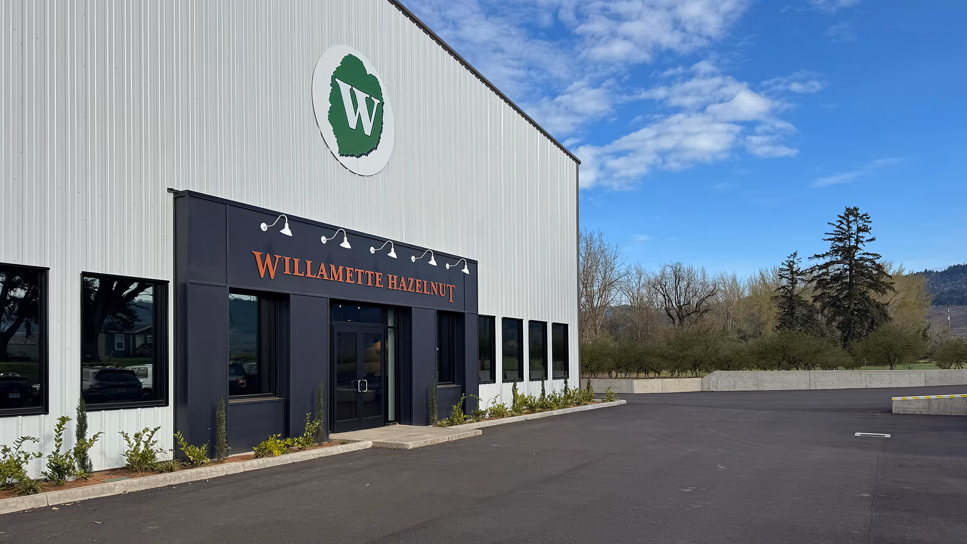 Entrance of a white industrial building with black doors and windows, labeled Willamette Hazelnut, under a partly cloudy blue sky.