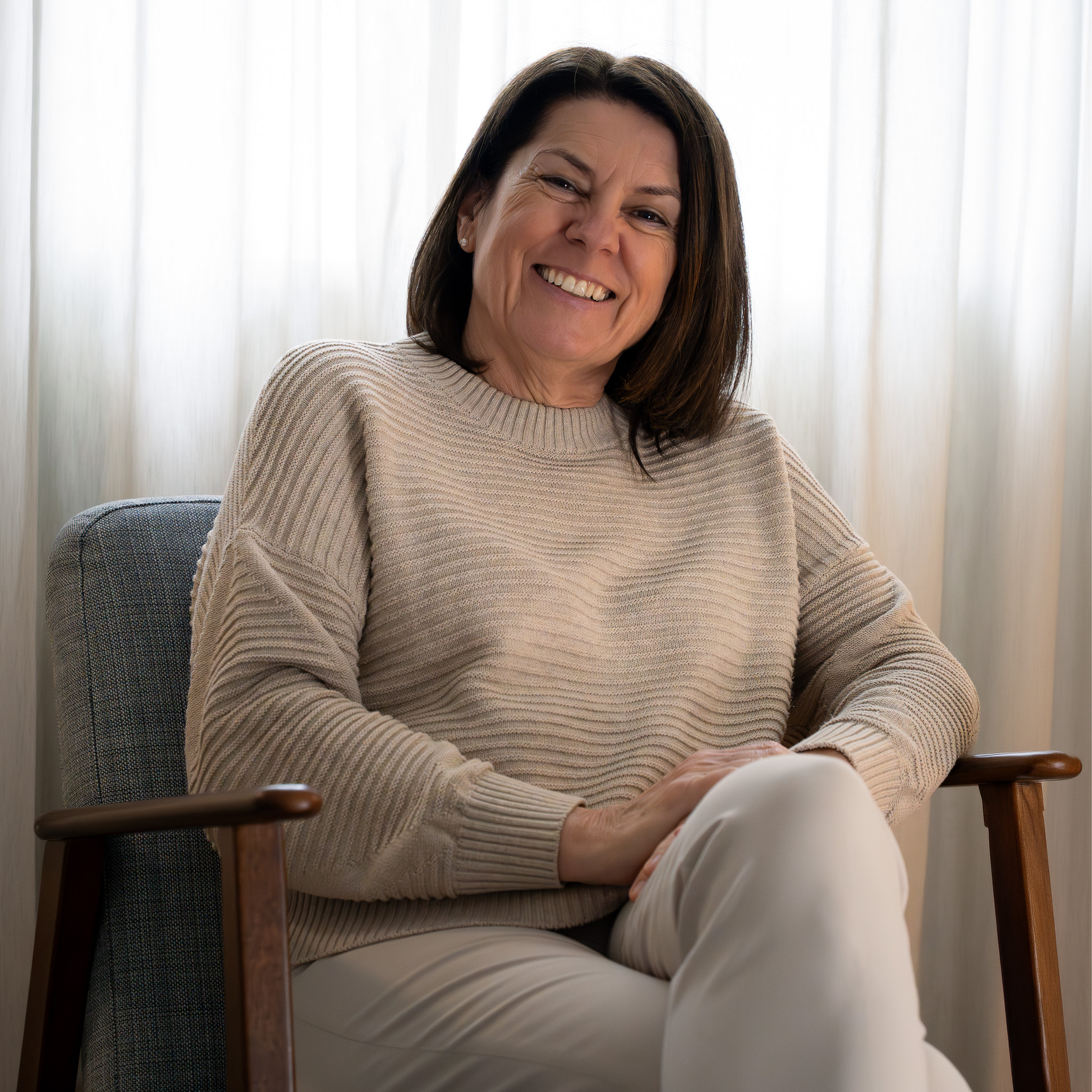 Lächelnde Frau mittleren Alters in beige gestricktem Pullover sitzt entspannt in einem Stuhl vor weißen Vorhängen.