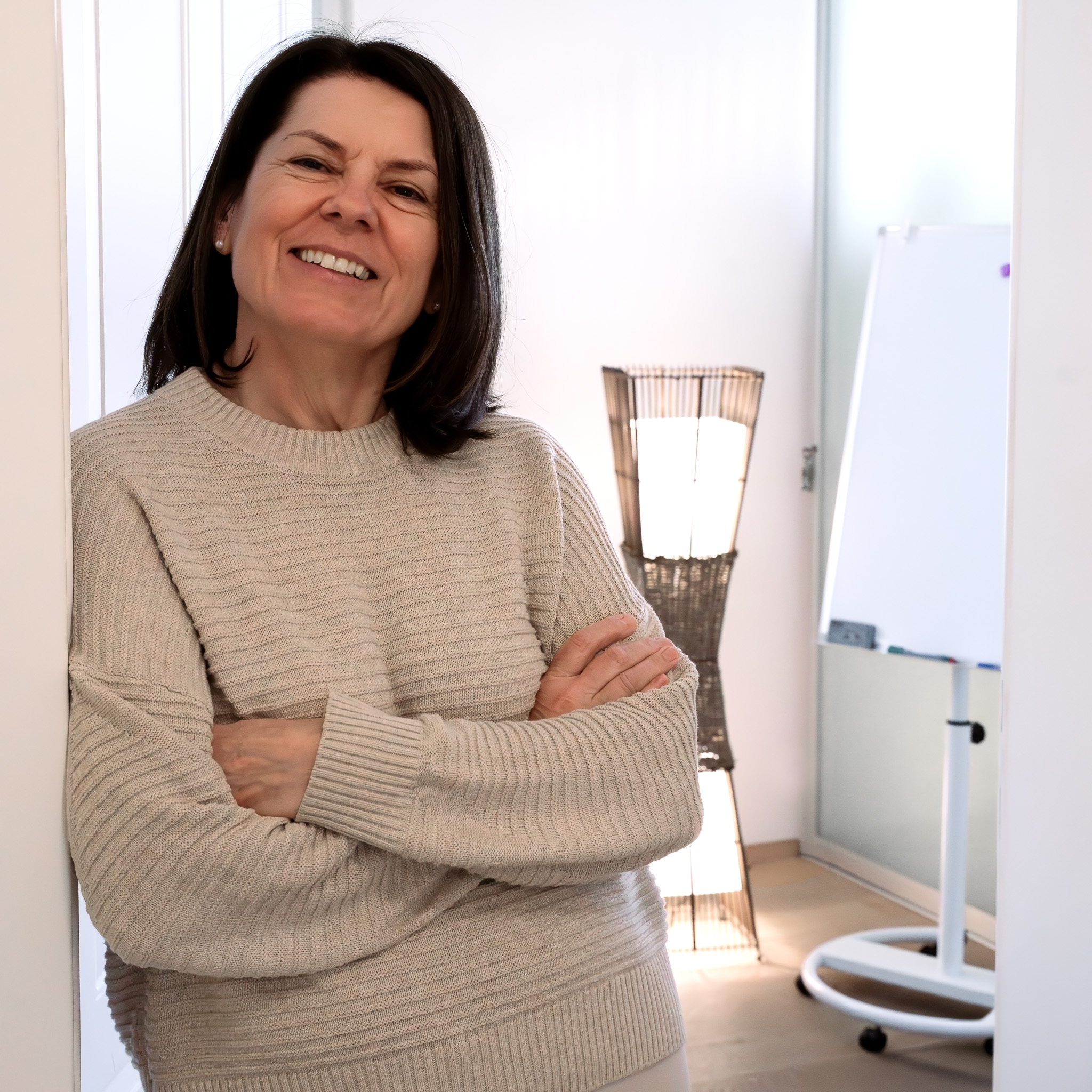 Lächelnde Frau mit beigem Pullover lehnt an einer Wand in einem hellen Raum mit Stehlampe und Whiteboard im Hintergrund.