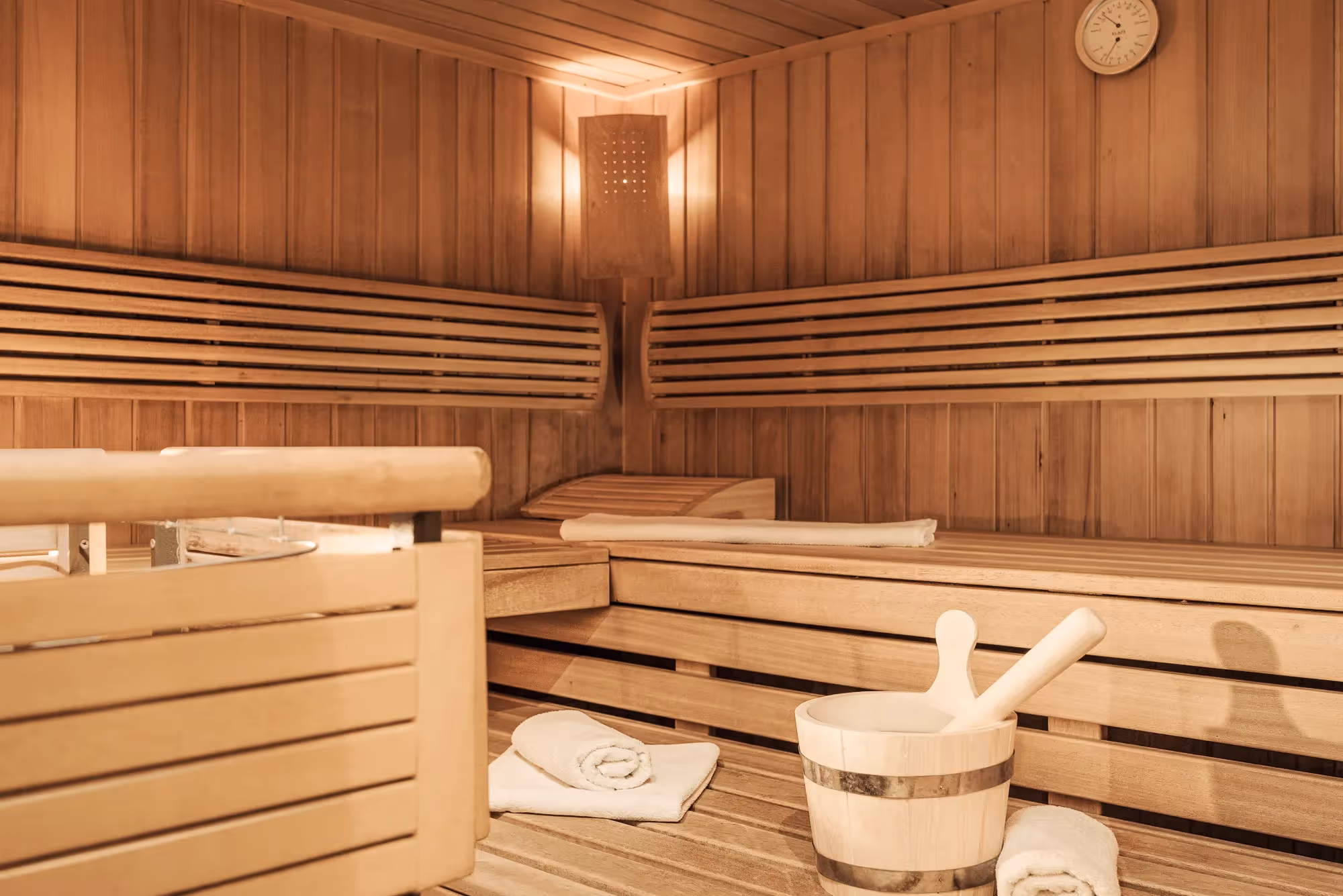 Innenraum einer Sauna mit Holzverkleidung, Holzbänken, Handtüchern und einem Holzeimer mit Schöpflöffel.