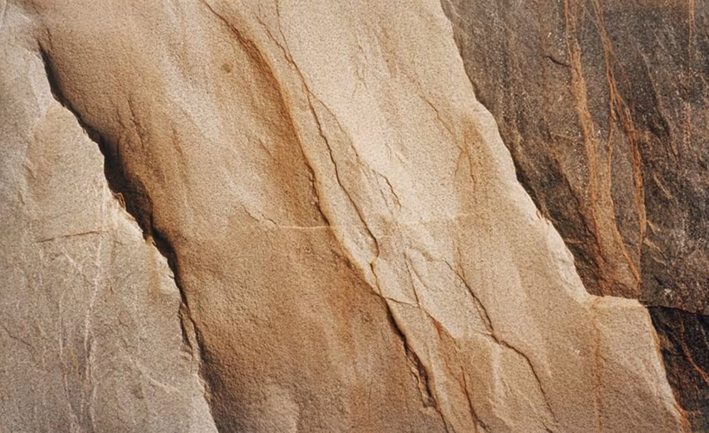 Close-up of a textured stone wall with various shades of beige, brown, and gray and visible cracks.