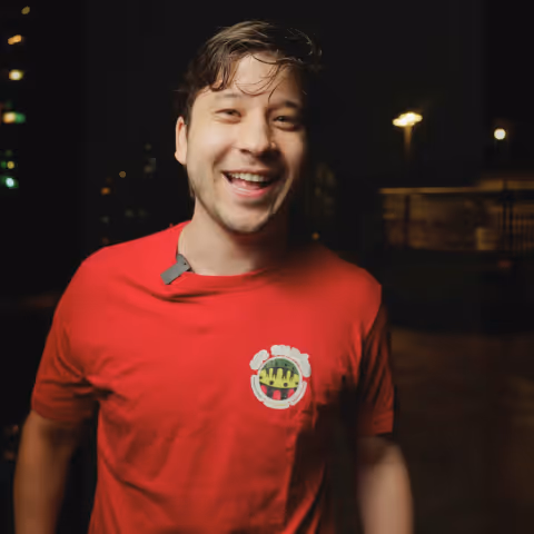 Smiling young man wearing a red t-shirt with a logo, standing outdoors at night.