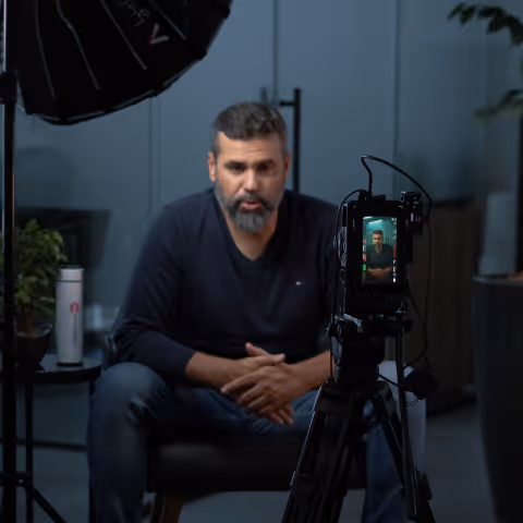 Man with a beard sitting in a chair being recorded by a camera on a tripod in a dimly lit room.