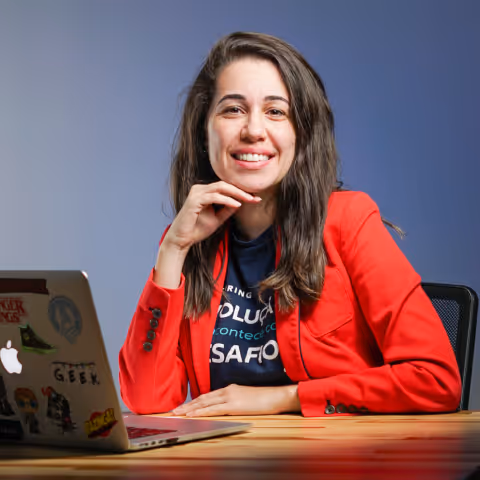 Smiling woman in a red blazer sitting at a table with a laptop covered in stickers against a blue background.