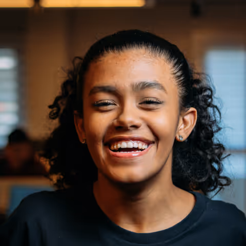 Young woman with curly hair smiling brightly indoors with blurred background.