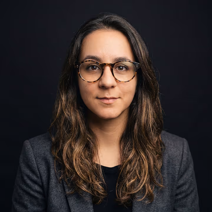 Woman with long brown wavy hair wearing round tortoiseshell glasses and a gray blazer against a dark background.