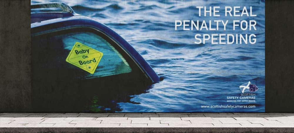 Partially submerged car in water with a 'Baby On Board' sign on the window and text warning 'The real penalty for speeding'.