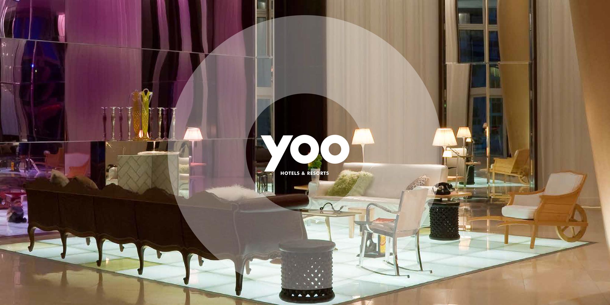 Modern hotel lobby with white sofa, wooden chairs, glass table, lamps, and reflective walls.