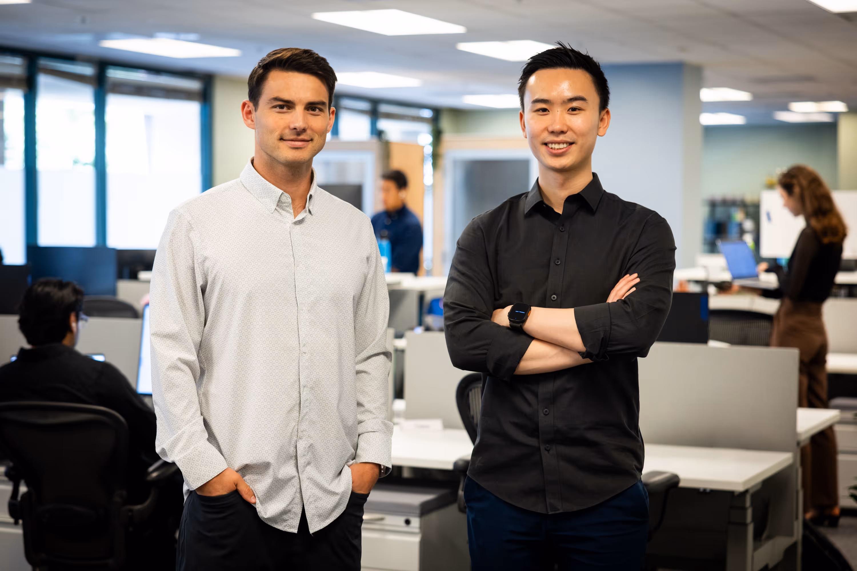 Two young men standing confidently in a modern office environment with colleagues working in the background.