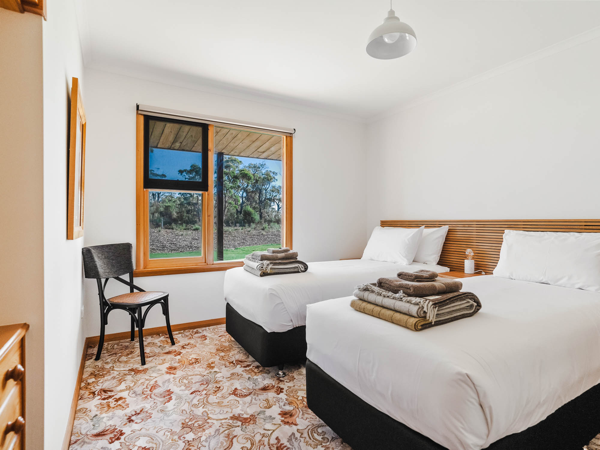 Bright bedroom with two single beds, white bedding, folded towels, a black chair, and a large window overlooking trees.