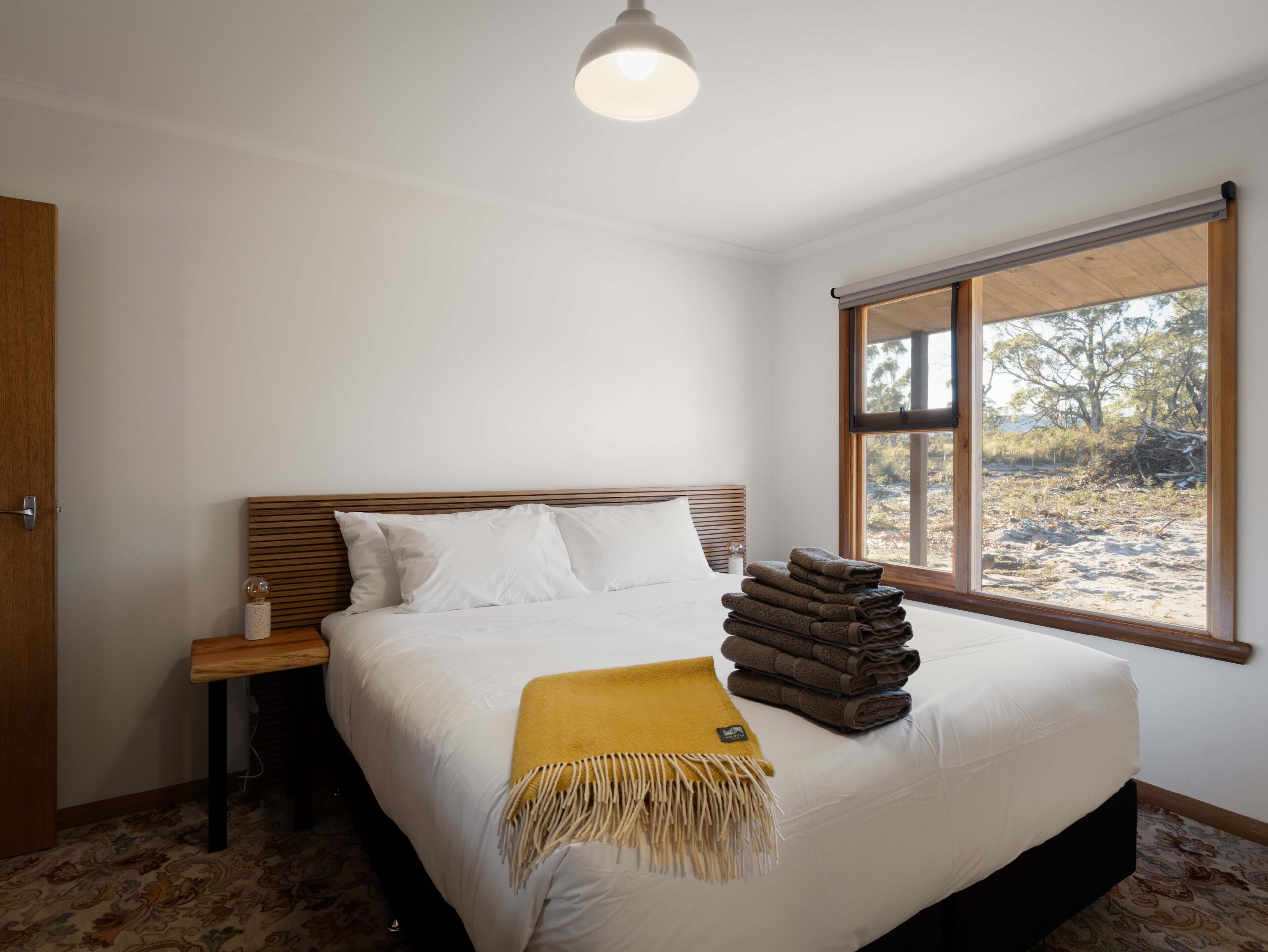 Minimalist bedroom with a large bed featuring white linens, a mustard yellow throw, a stack of brown towels, wooden headboard, window showing outdoor natural scenery.