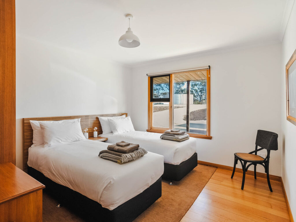 Bright bedroom with two single beds, white linens, folded towels on each bed, wooden floor, window with a view, and a black chair.