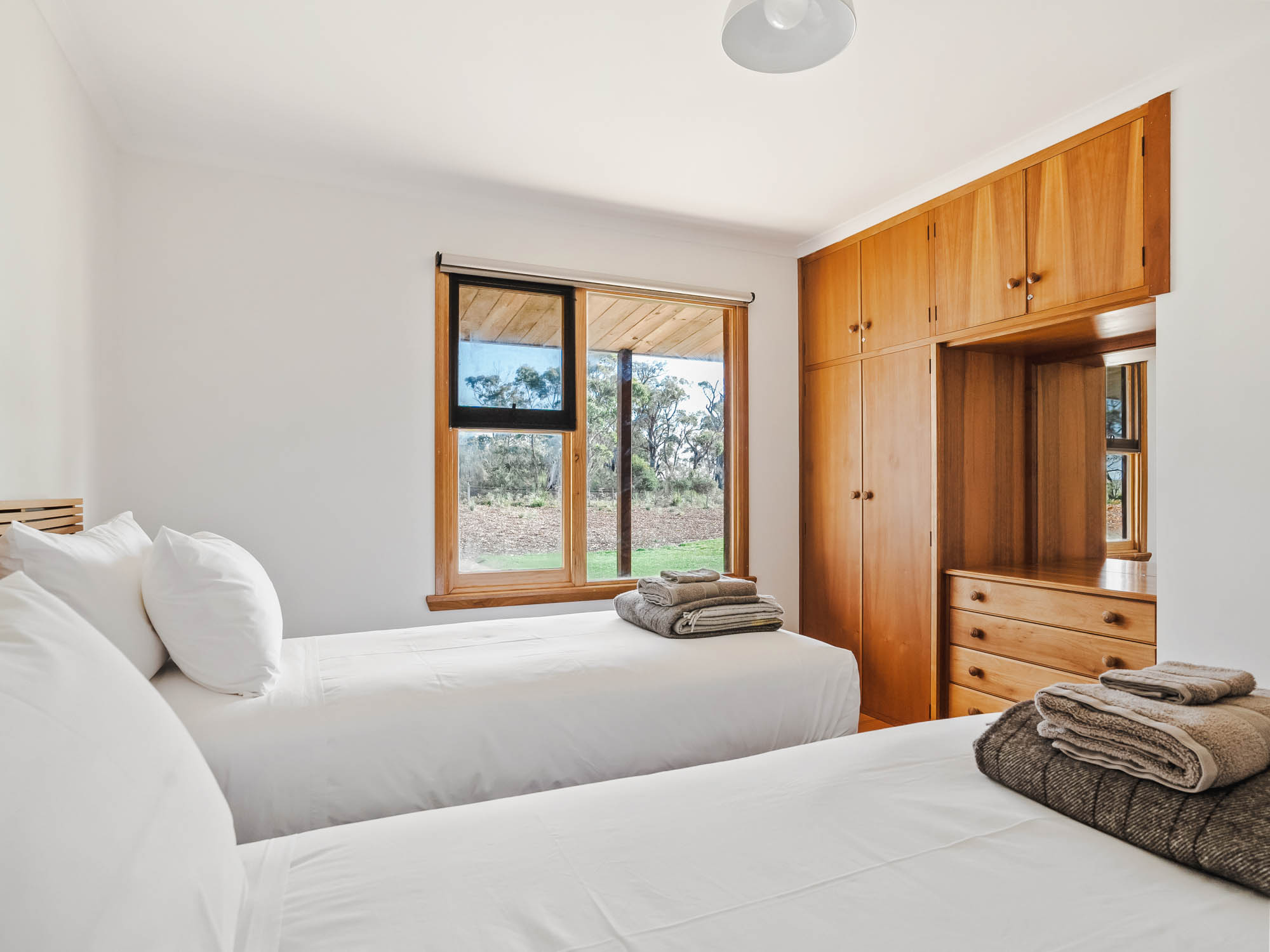 Minimalist bedroom with two single beds, white linens, wooden wardrobes, and a window showing an outdoor landscape.
