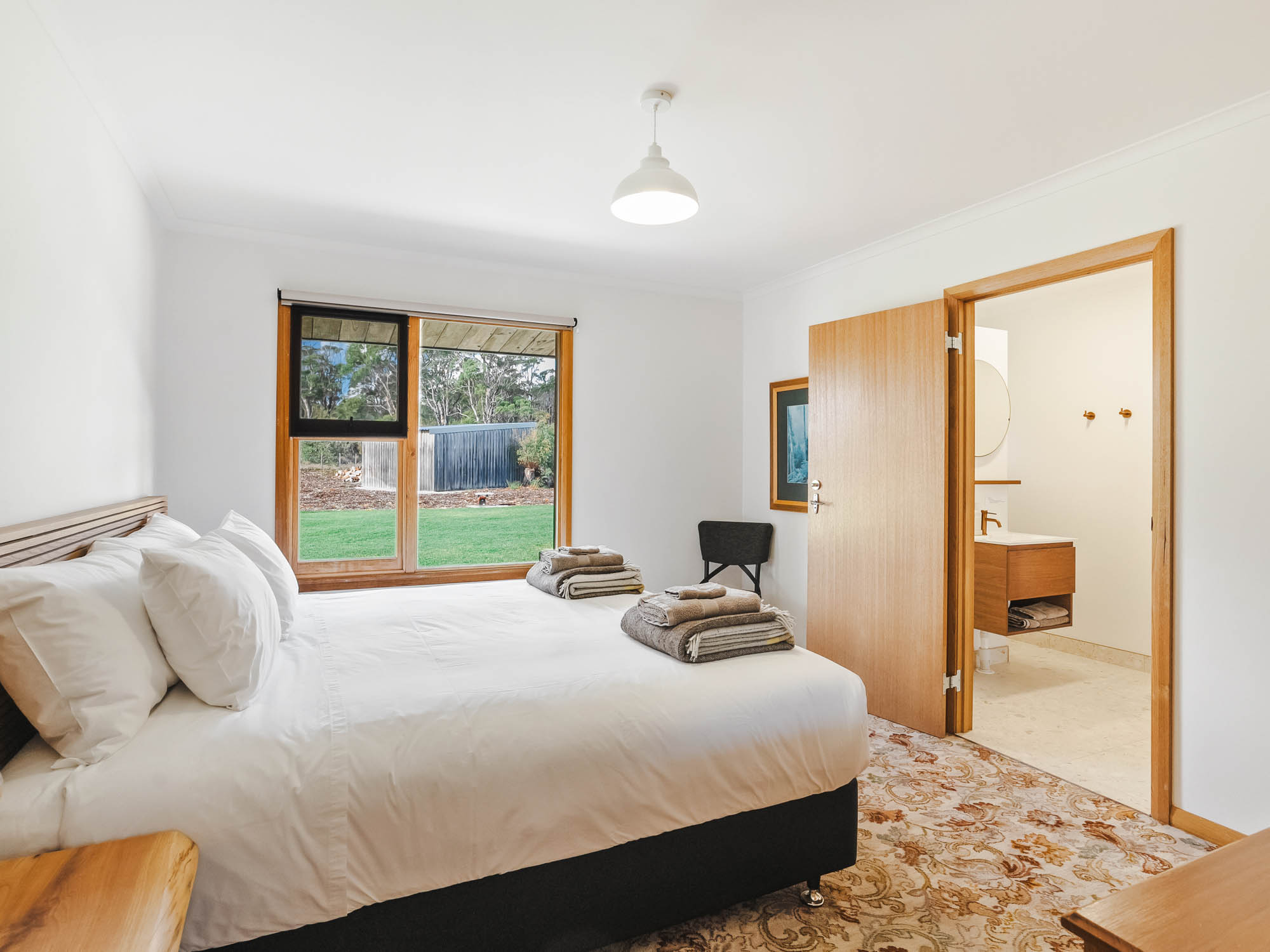 Bright bedroom with large window overlooking green yard, white bedding, folded towels on bed, and an open door to a bathroom.