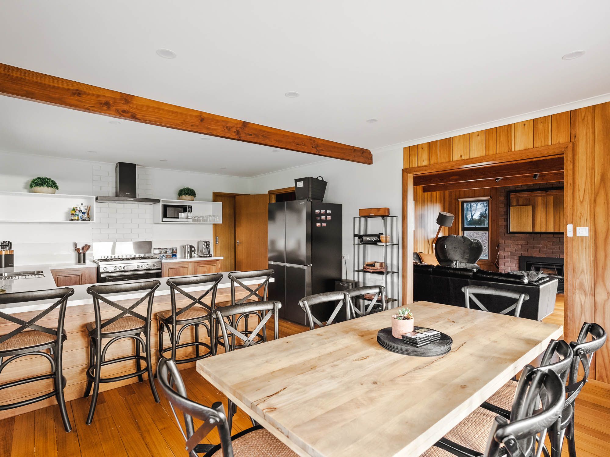Modern kitchen and dining area with wooden floor, black metal chairs, wooden dining table, black refrigerator, and wood-paneled living room visible through an open doorway.