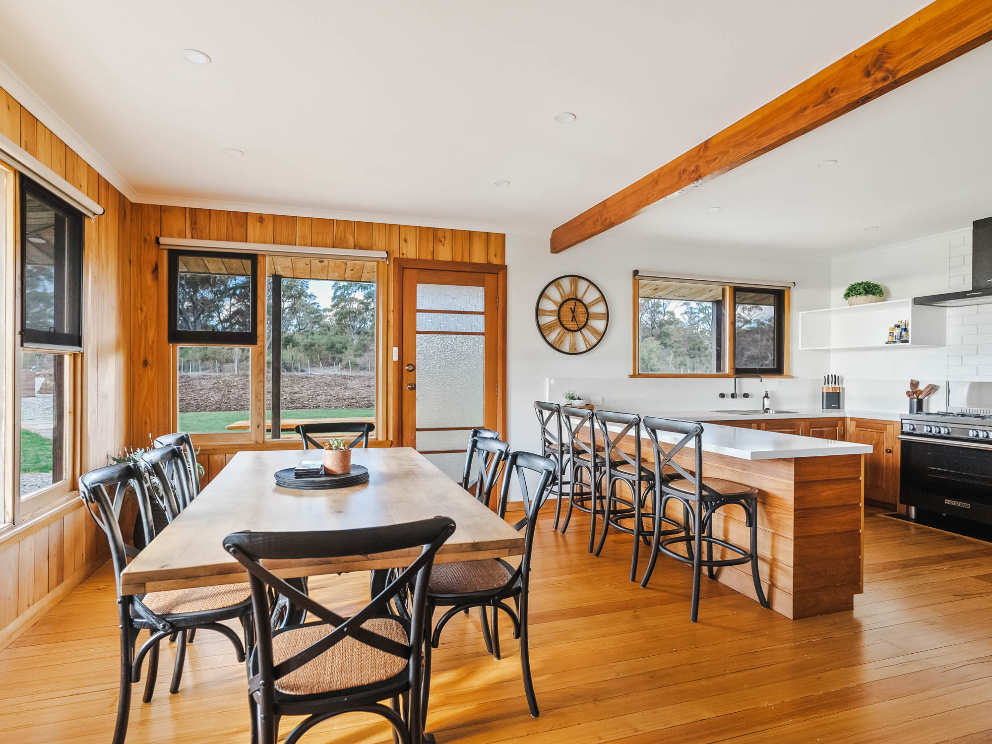 Bright kitchen and dining area with wood paneling, a large wooden table with black chairs, a kitchen island with stools, and large windows showing outdoor trees.