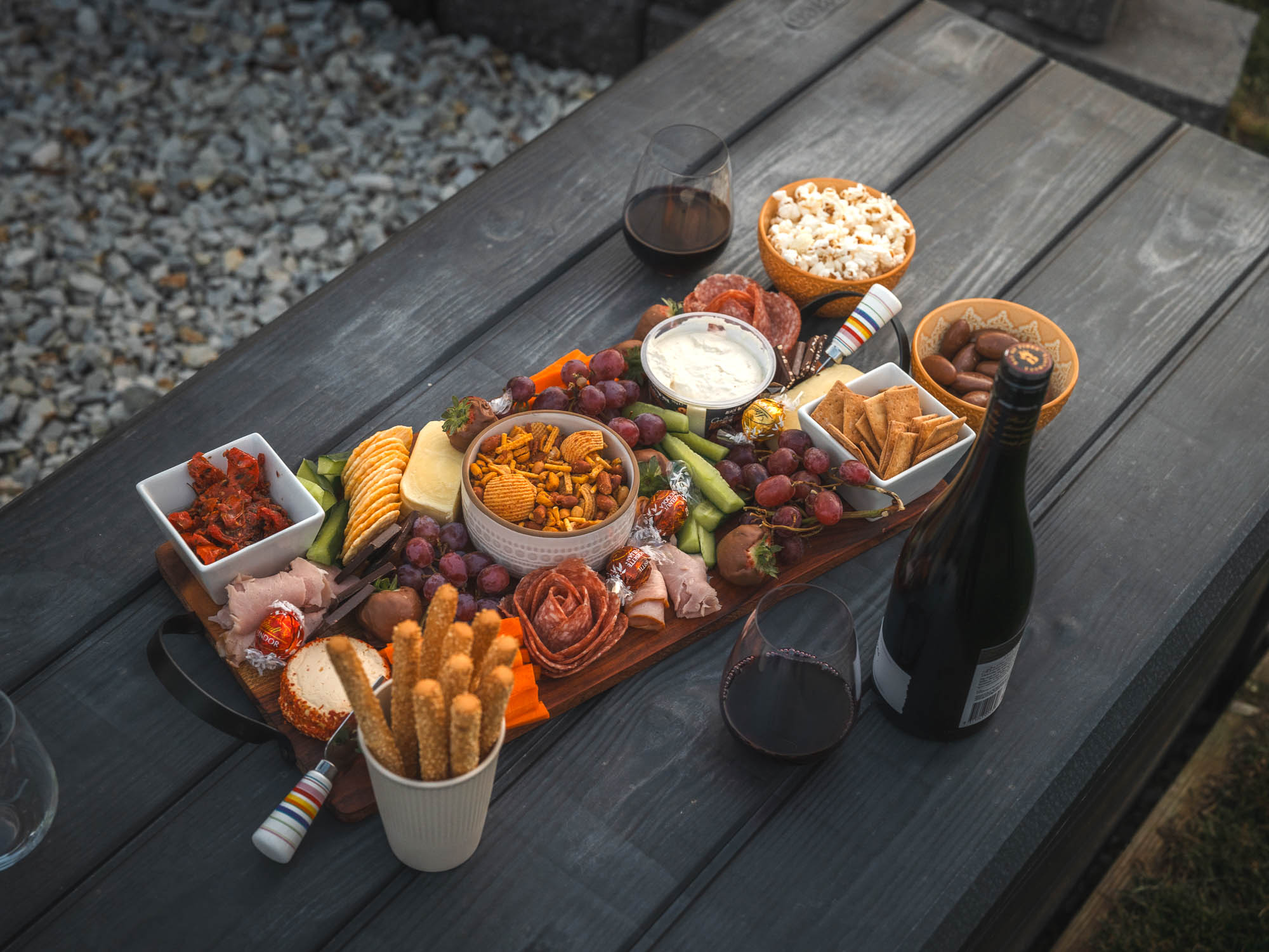 Charcuterie board with snacks including grapes, crackers, cheese, meats, dips, popcorn, and breadsticks beside a bottle and glasses of red wine on a dark picnic table.