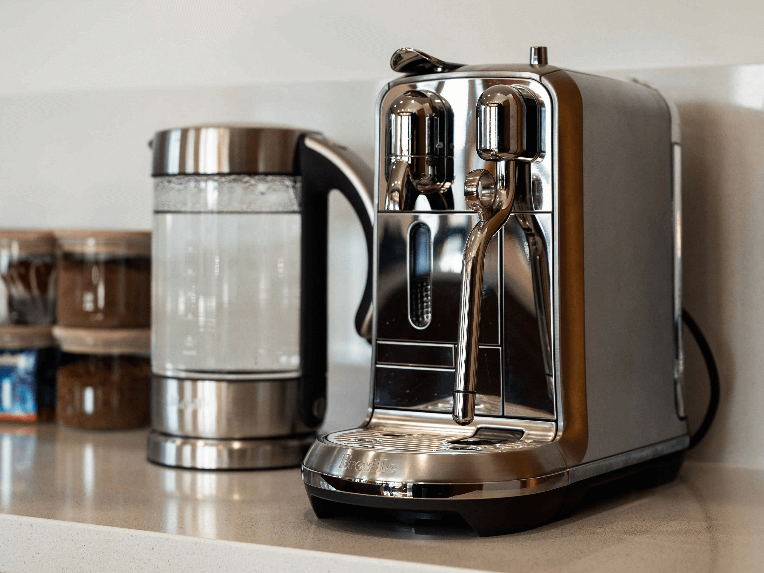 Stainless steel espresso machine and electric kettle on a kitchen countertop with jars in the background.