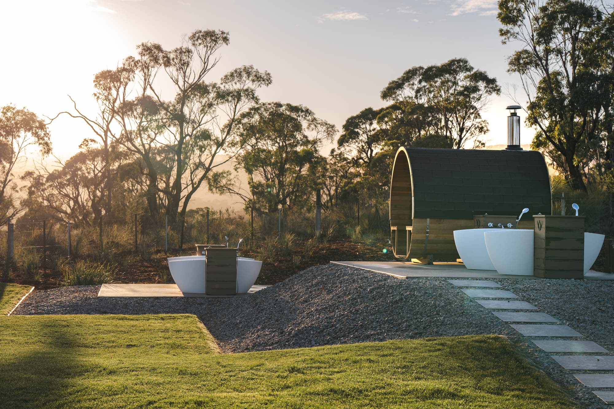 Outdoor setting with two white bathtubs and a wooden barrel sauna surrounded by trees and grass at sunset.