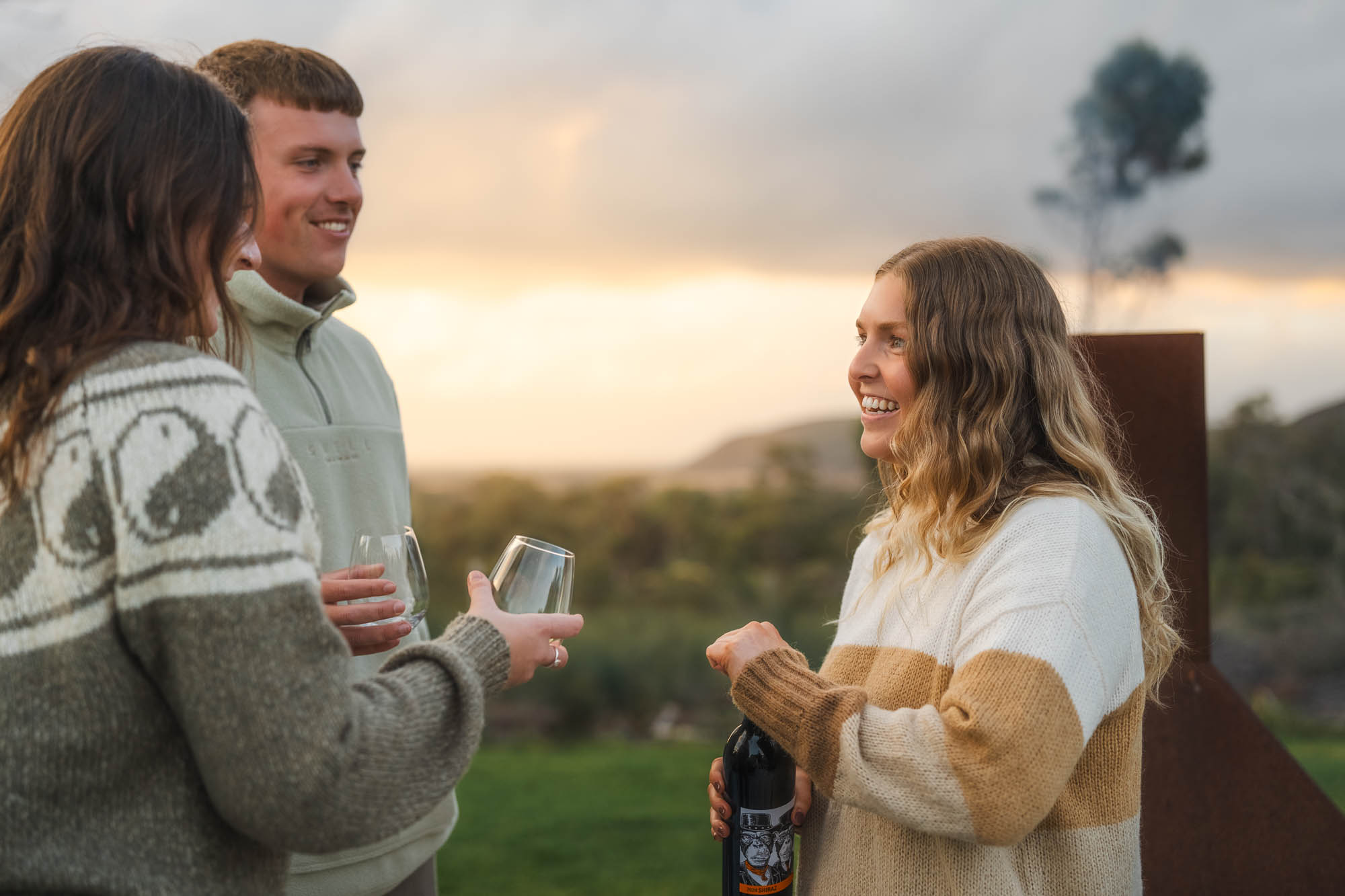 Three people outdoors at sunset enjoying wine, with one woman holding a wine bottle and two others holding empty wine glasses.