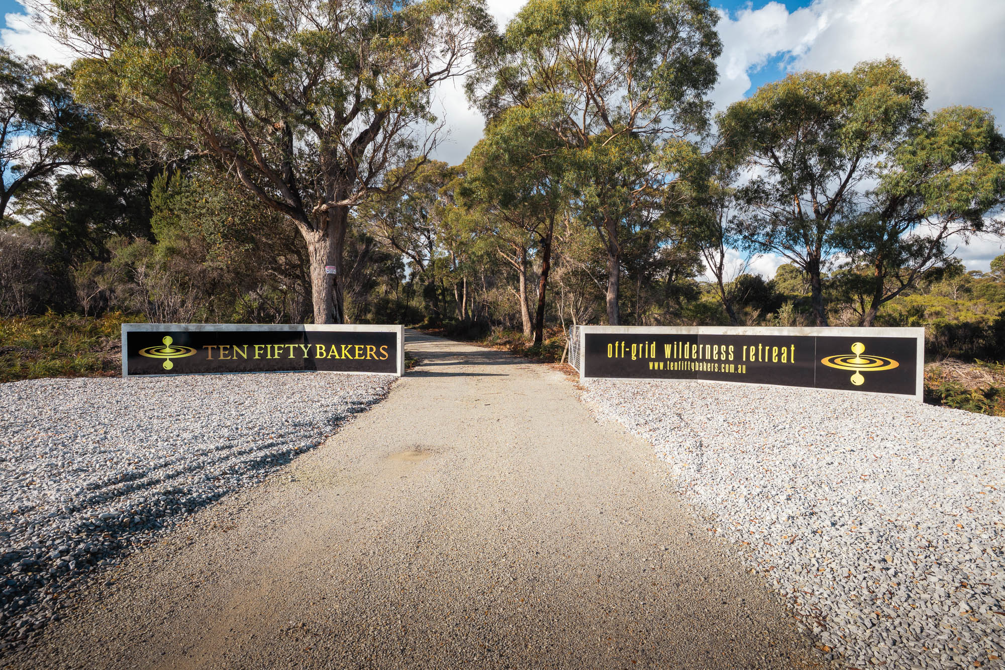 Gravel driveway entrance flanked by black signs with yellow text reading Ten Fifty Bakers and off-grid wilderness retreat surrounded by trees.