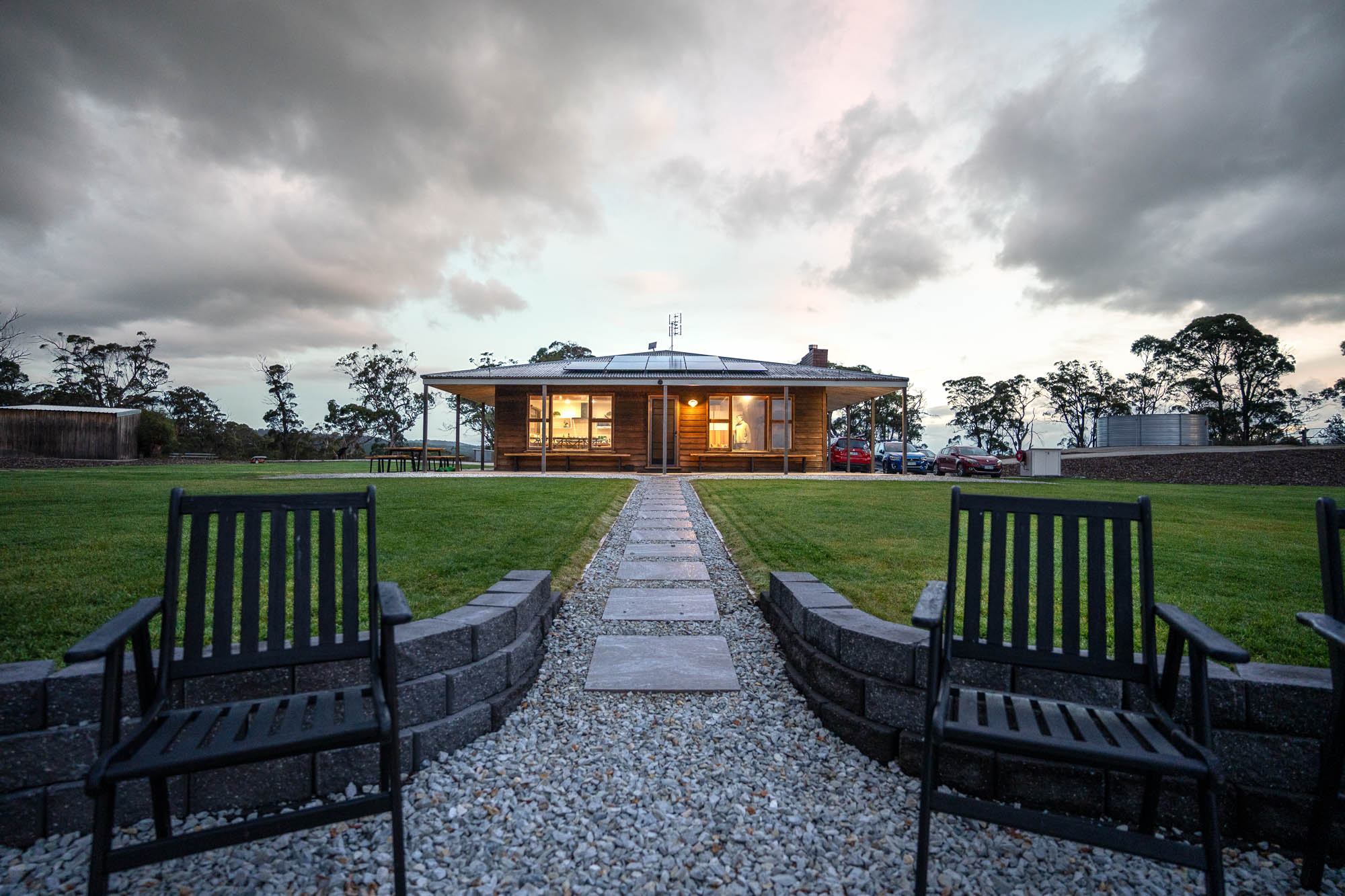 Path leading to a modern wooden house with lit windows and solar panels on the roof, flanked by green lawns and black outdoor chairs under a cloudy sky at dusk.