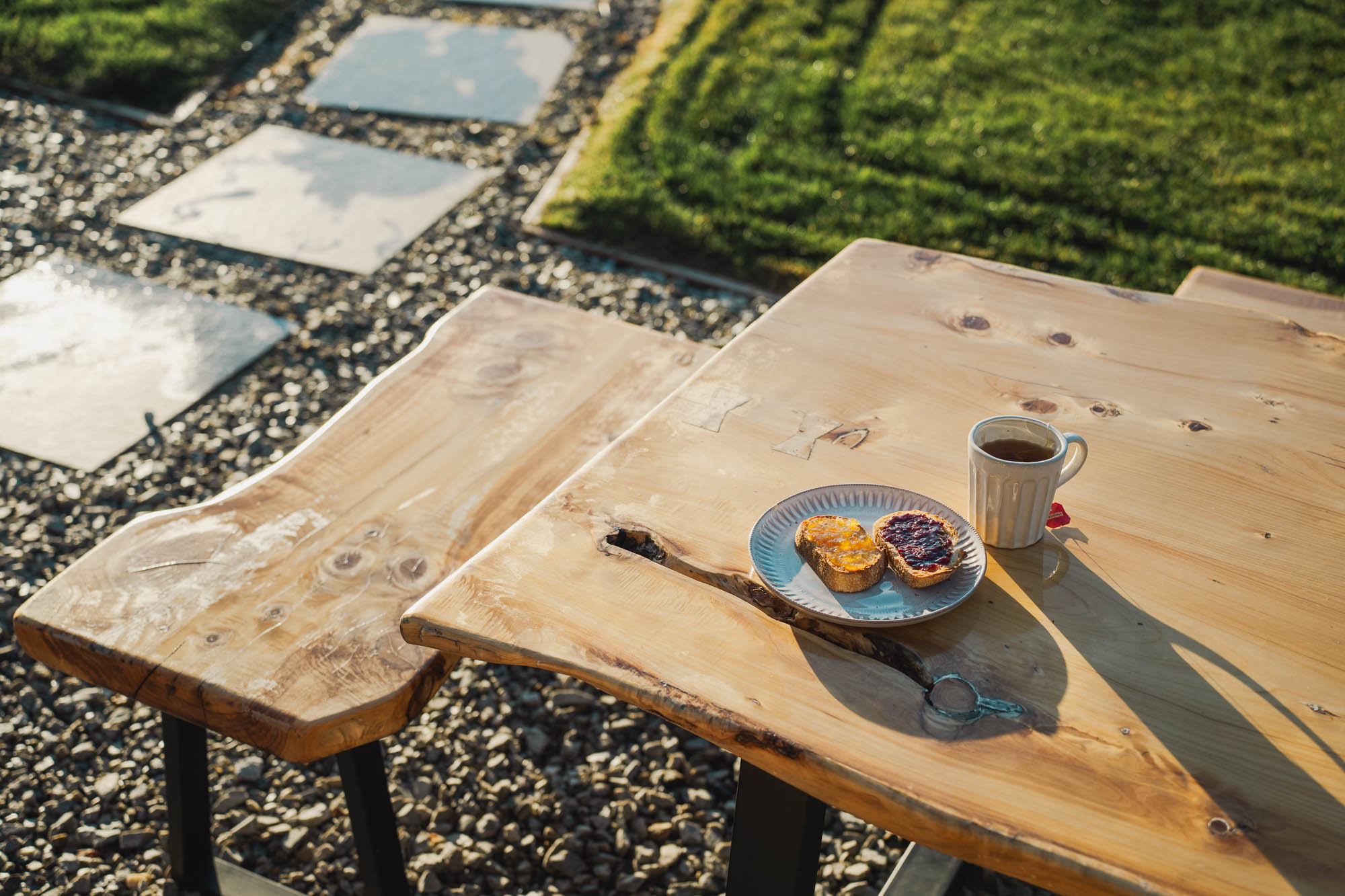 Wooden outdoor table and bench with a plate of toast spread with jam and marmalade, and a cup of tea.