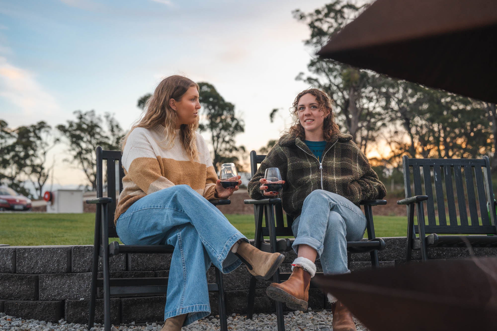 Two women sitting outdoors in chairs, drinking red wine and talking around a fire pit at sunset.
