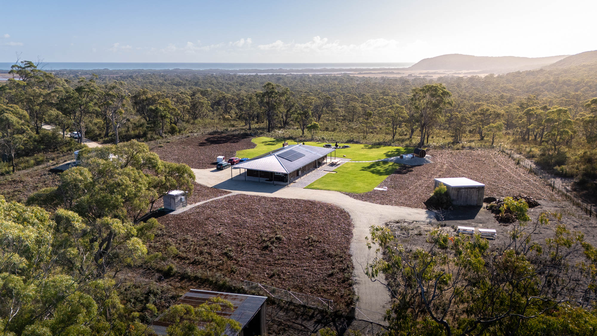 Aerial view of a single-story house with solar panels on the roof, surrounded by green lawn, dirt paths, scattered buildings, and dense forest extending to distant hills under a clear sky.