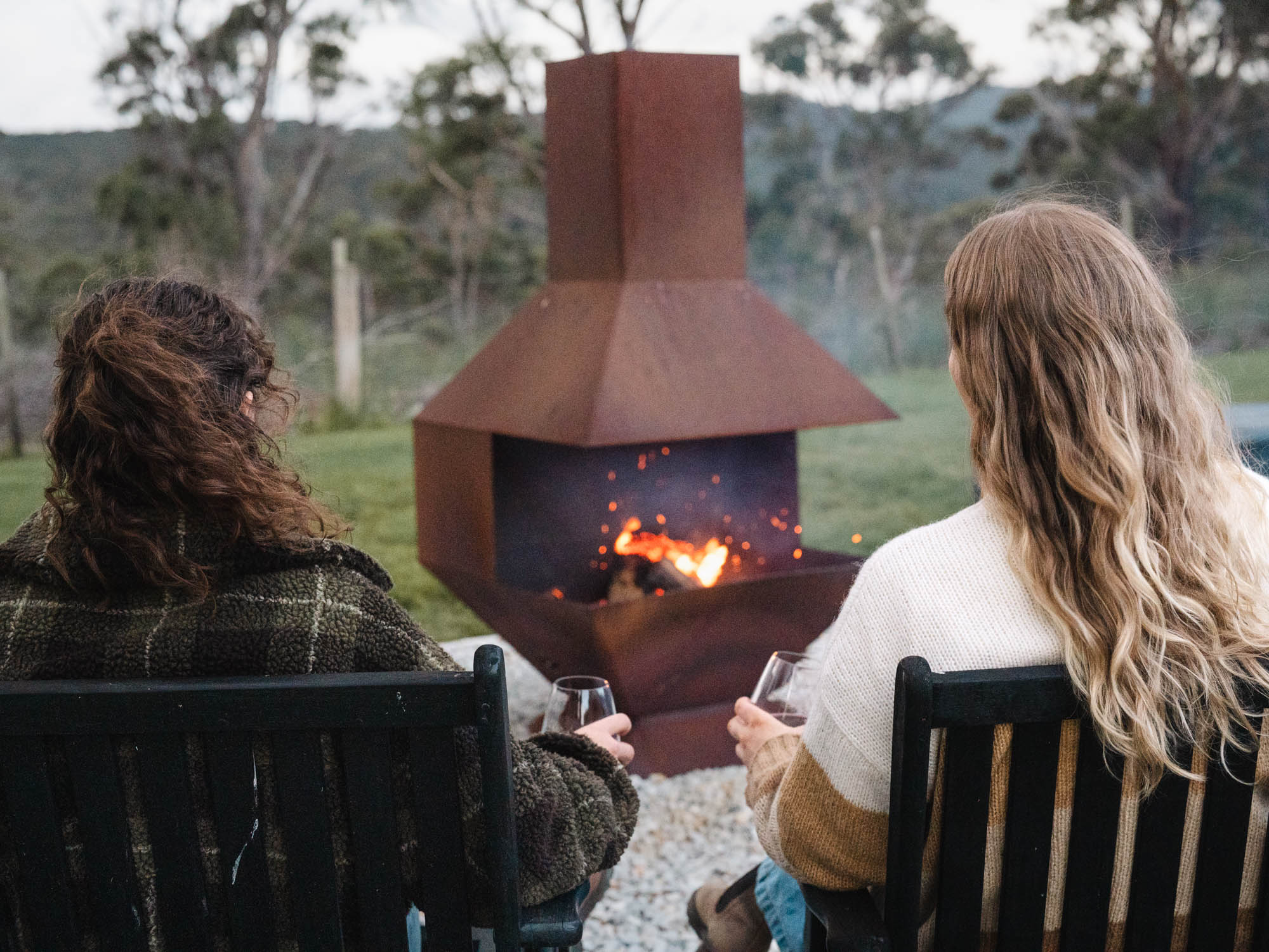 Two women sitting on chairs by an outdoor fire pit, each holding a glass of wine, surrounded by green grass and trees.