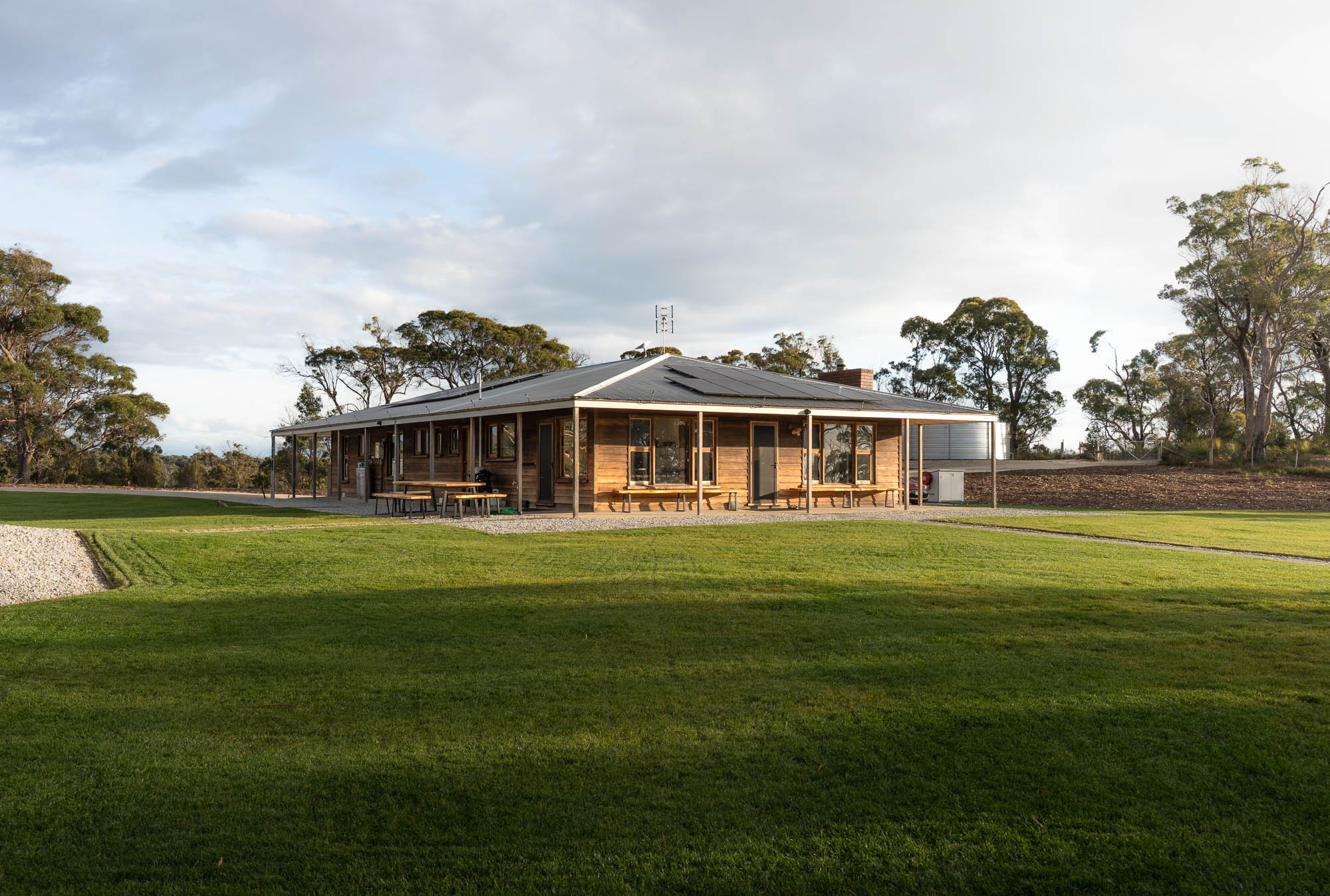 Single-story wooden house with solar panels on the roof, surrounded by green grass and trees under a cloudy sky.
