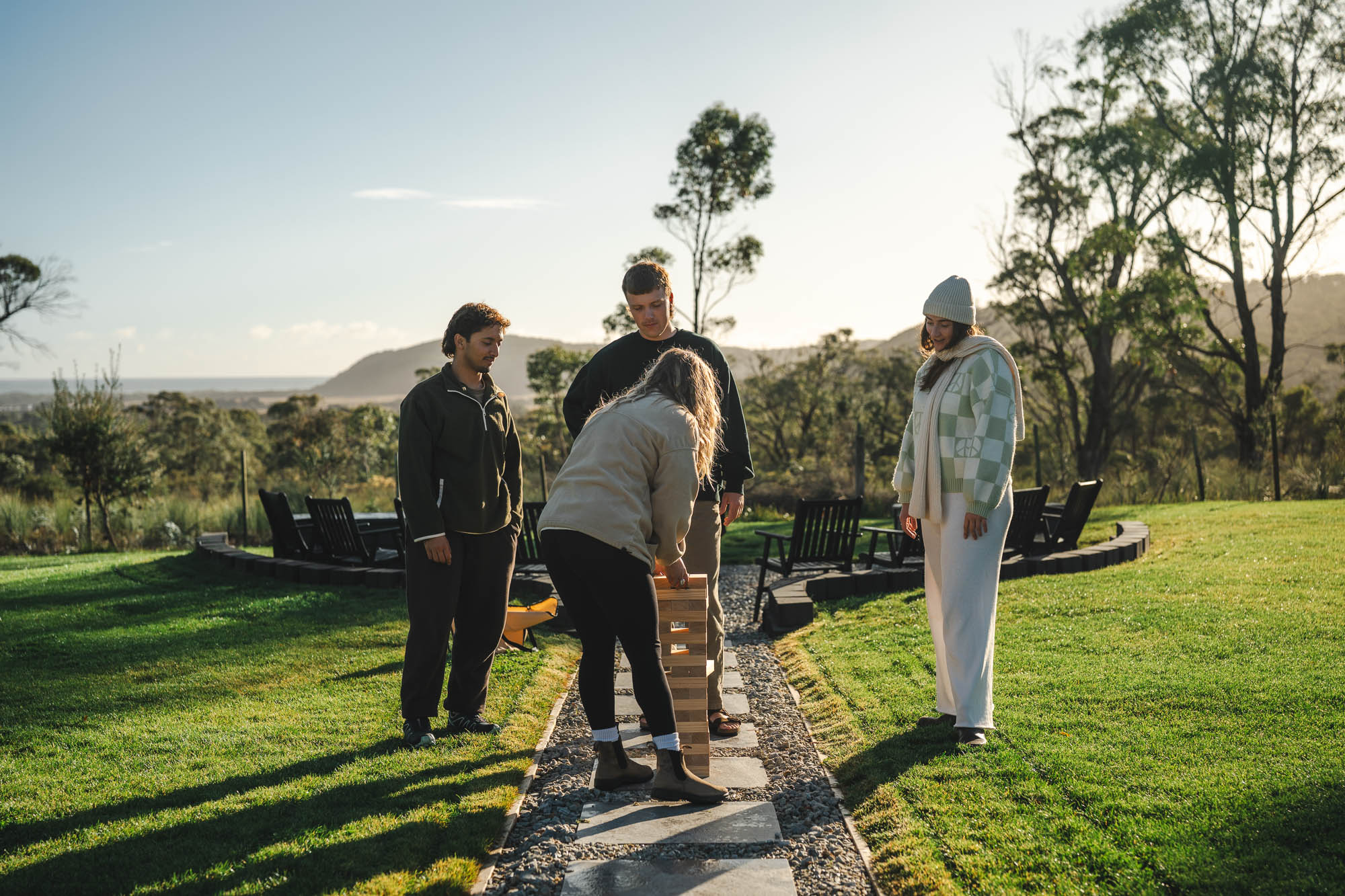 Four people playing a large outdoor Jenga game on a stone path surrounded by grass and trees.