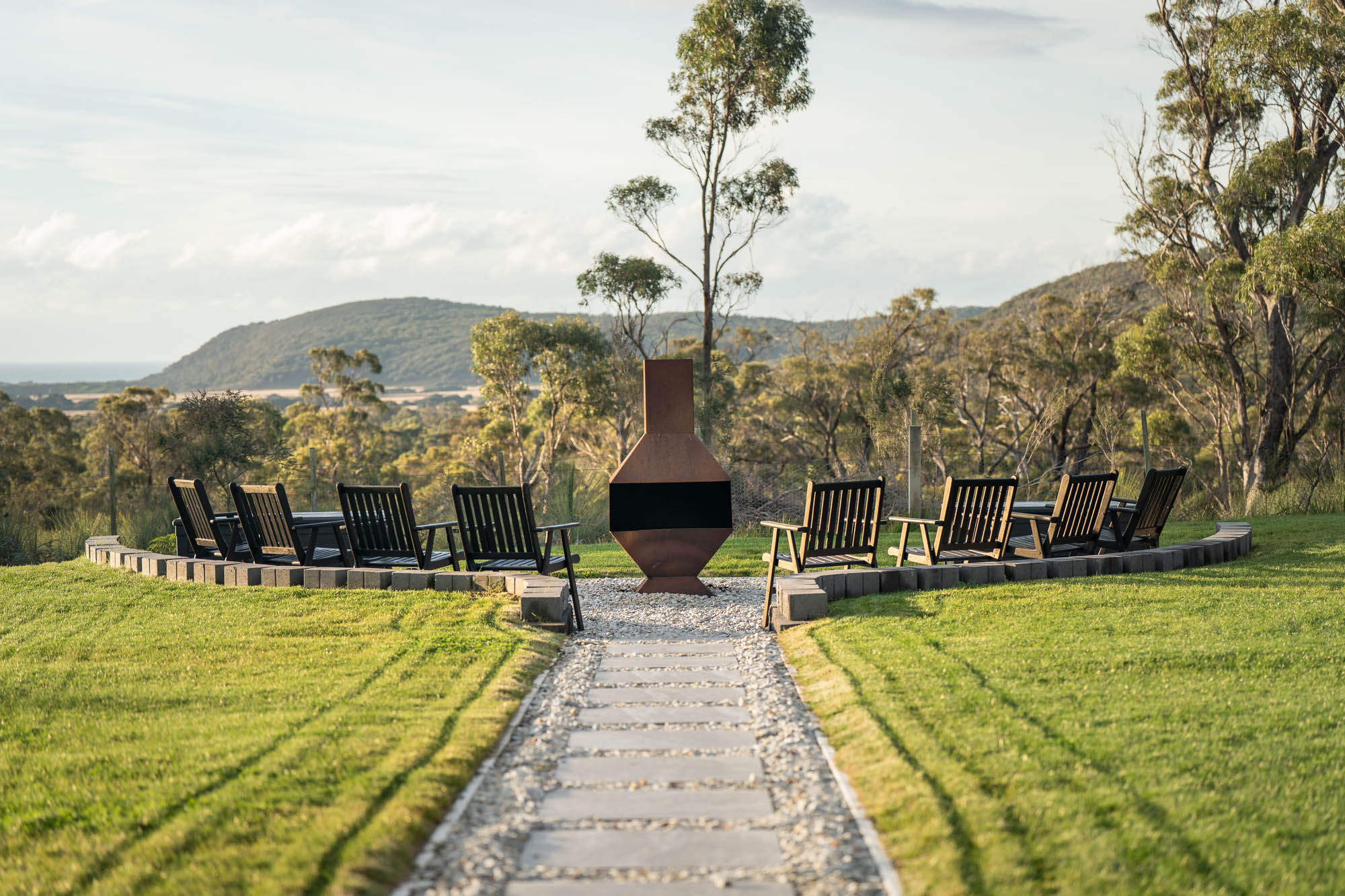 Outdoor seating area with wooden chairs arranged in a semicircle around a central metal fire pit on a grassy lawn with hills and trees in the background.