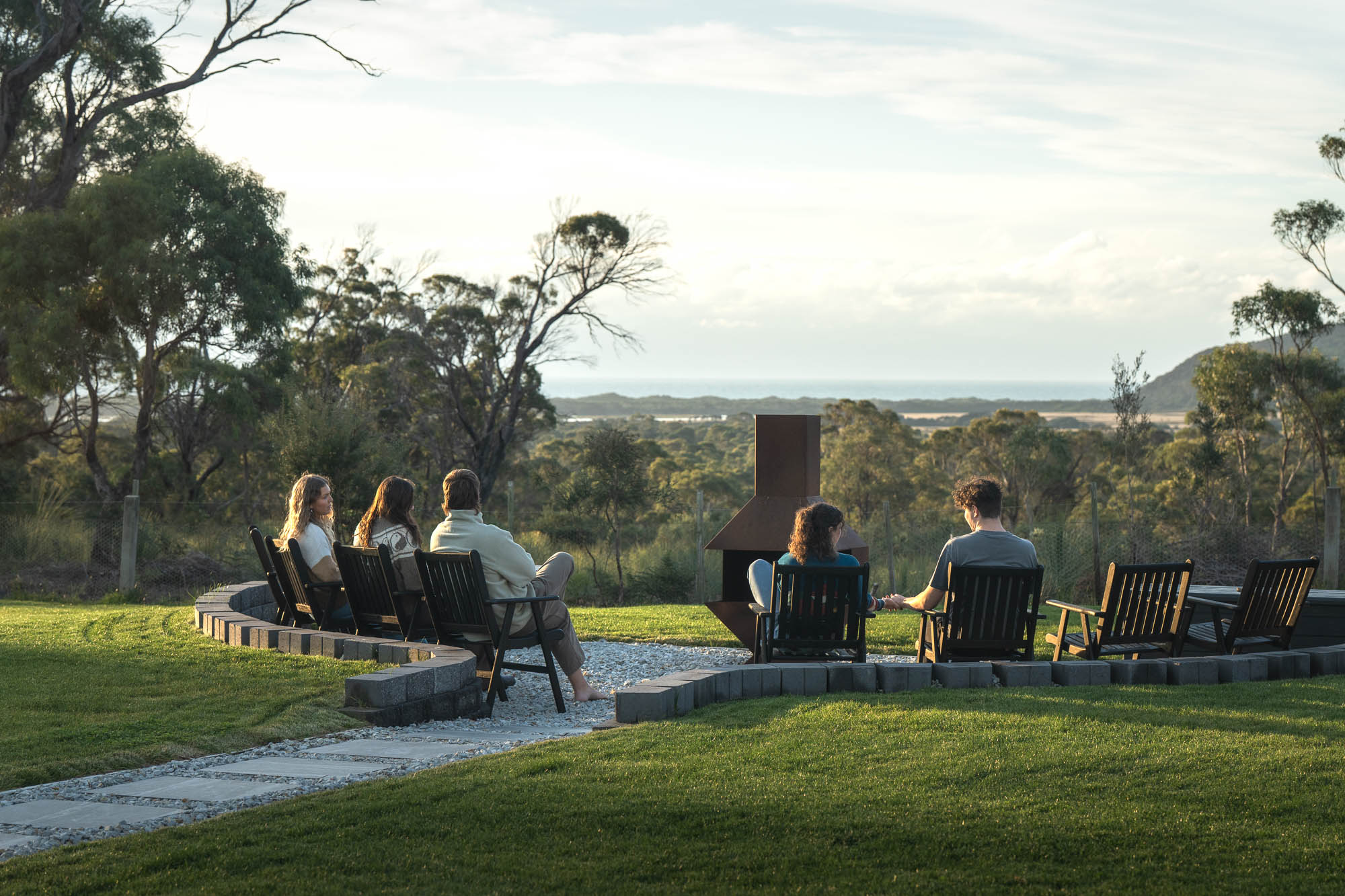 Five people sitting in wooden chairs around an outdoor fire pit in a grassy area, overlooking trees and distant hills under a cloudy sky.