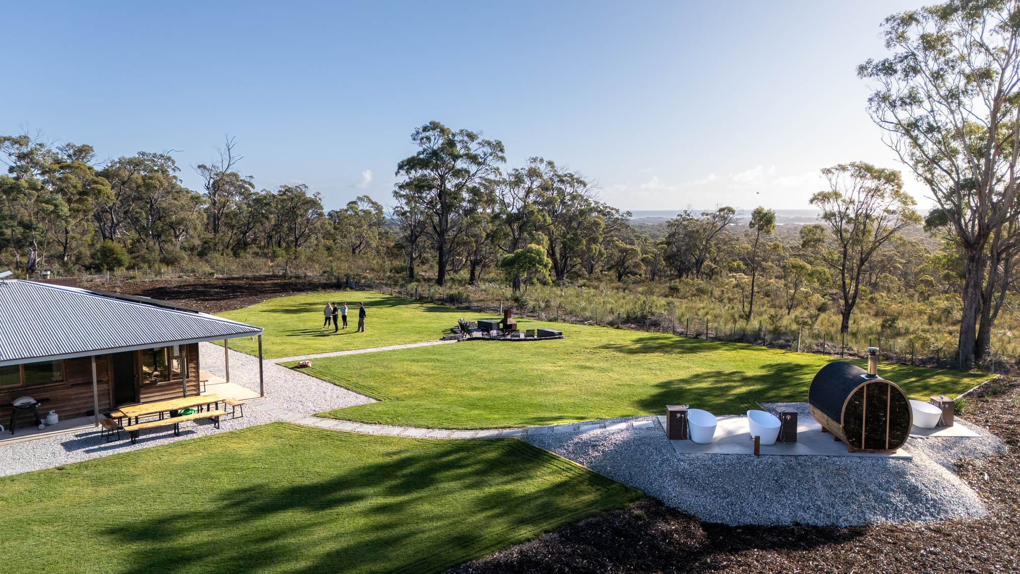 A group of people standing on a large green lawn near a wooden cabin with outdoor seating, surrounded by trees and nature.