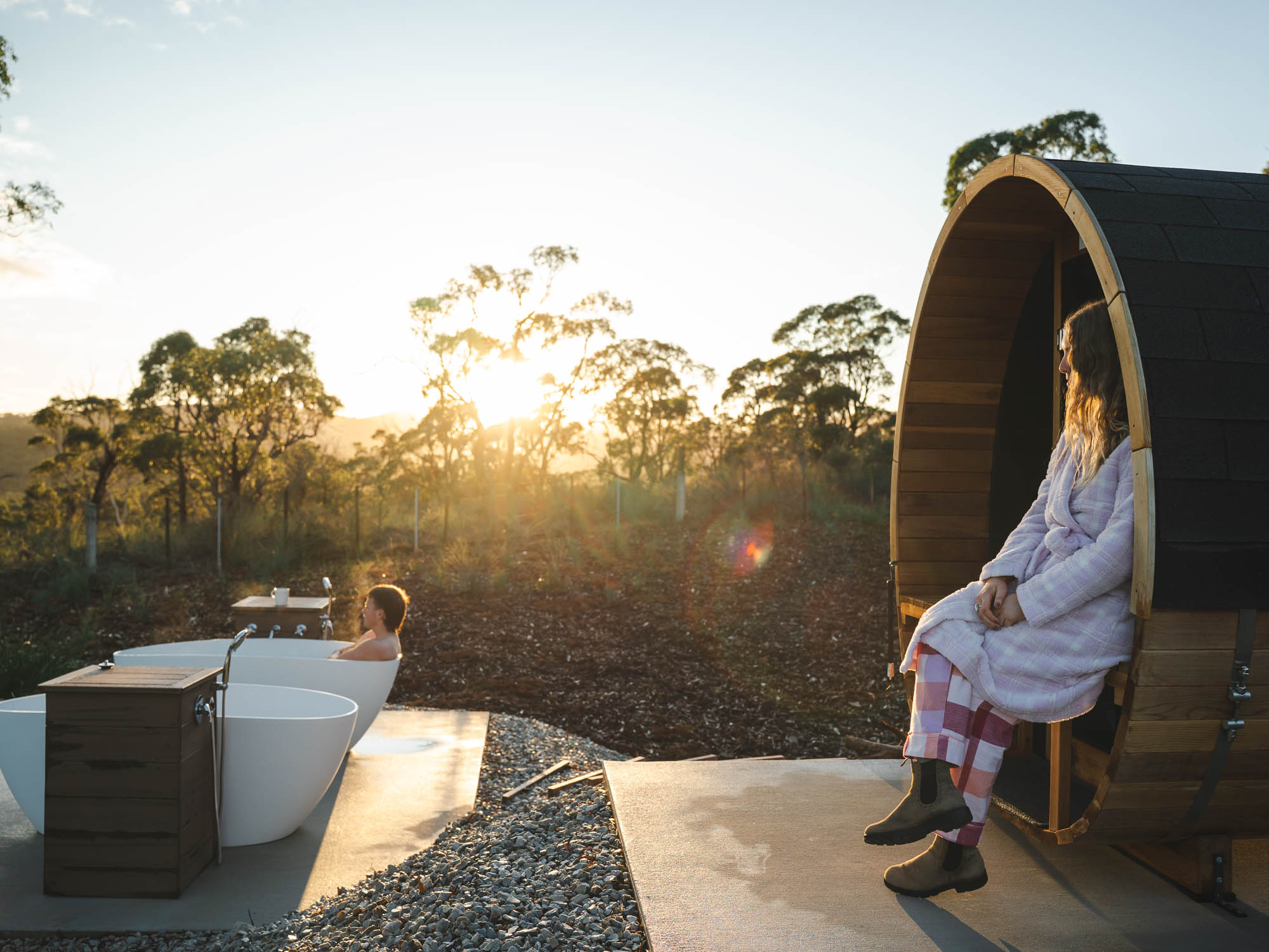Woman in a bathrobe sitting at the entrance of a wooden barrel sauna next to a person relaxing in an outdoor white bathtub at sunset.