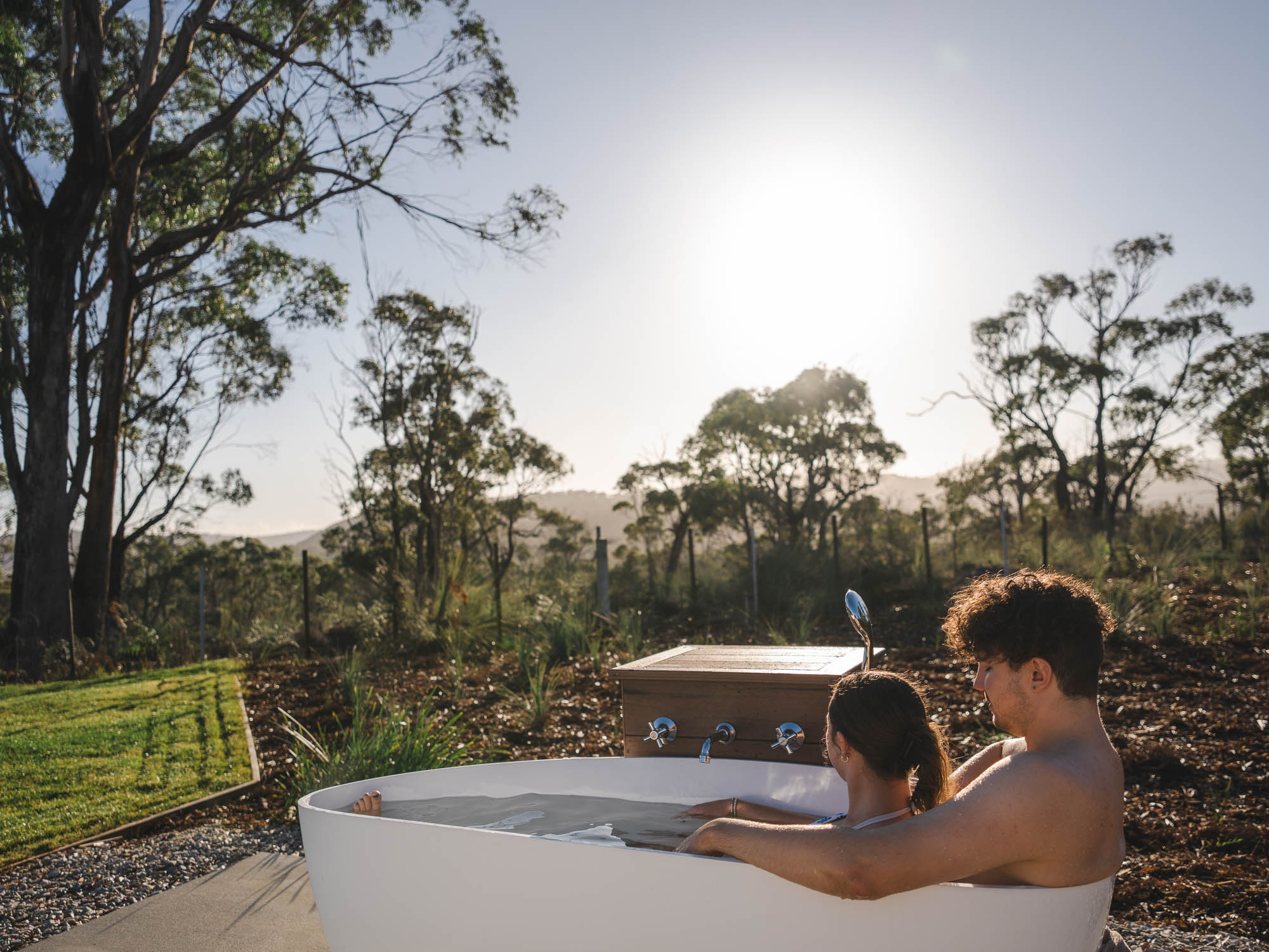 Young couple relaxing together in an outdoor bathtub at sunset surrounded by trees and nature.