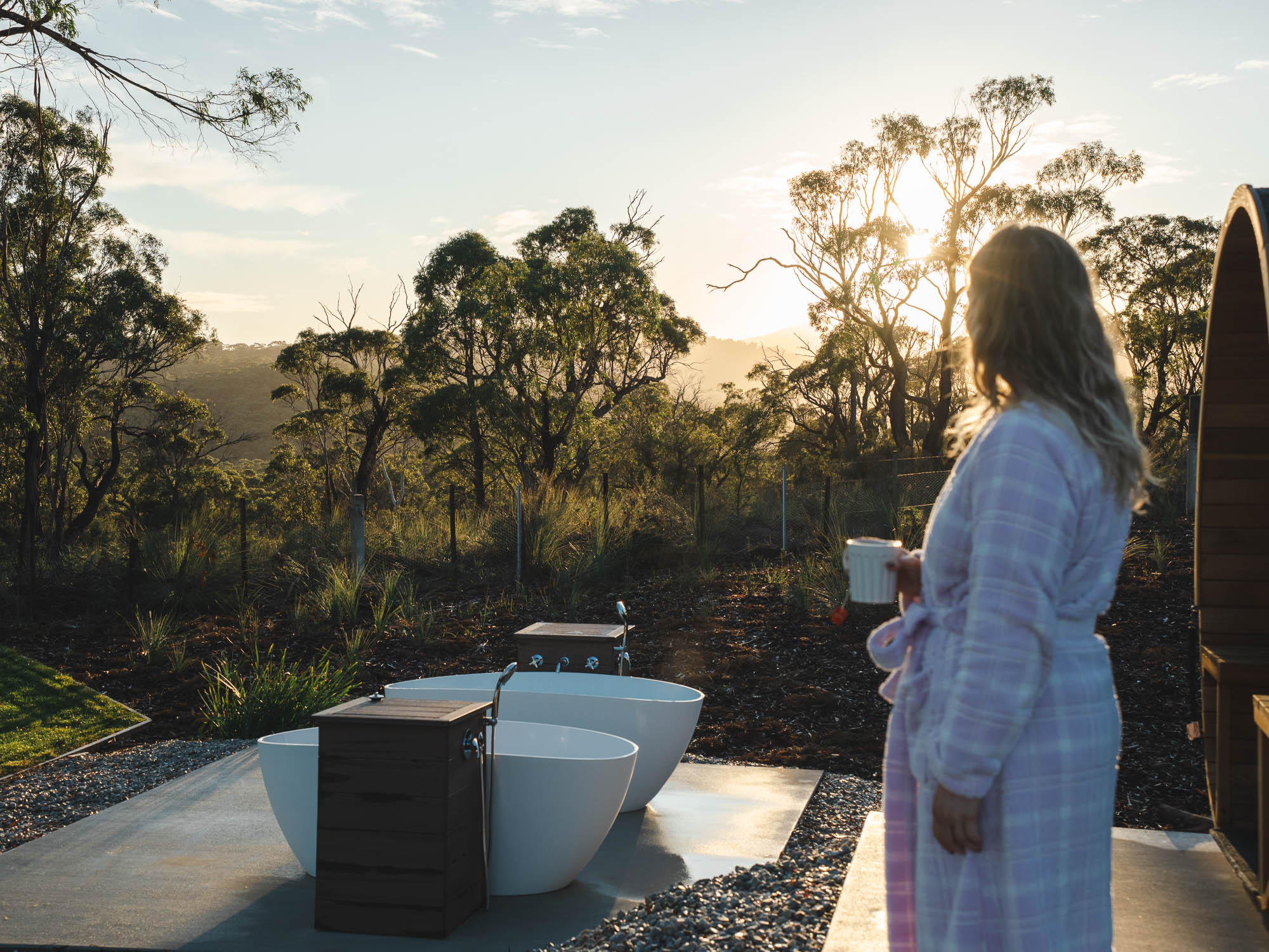 Woman in a lavender robe holding a cup, looking towards two white outdoor bathtubs with wooden fixtures at sunset in a natural forest setting.