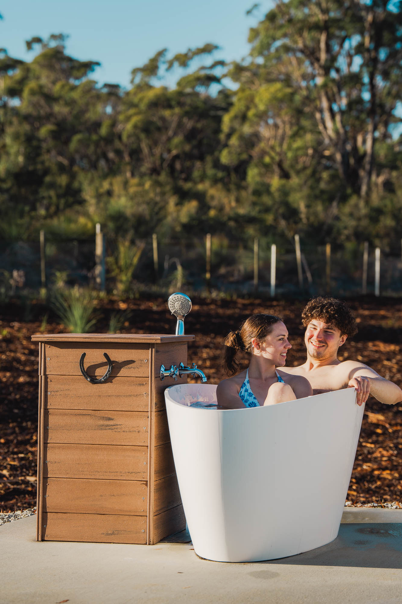 Couple smiling and sitting together in an outdoor white bathtub beside a wooden shower with a horseshoe decoration.