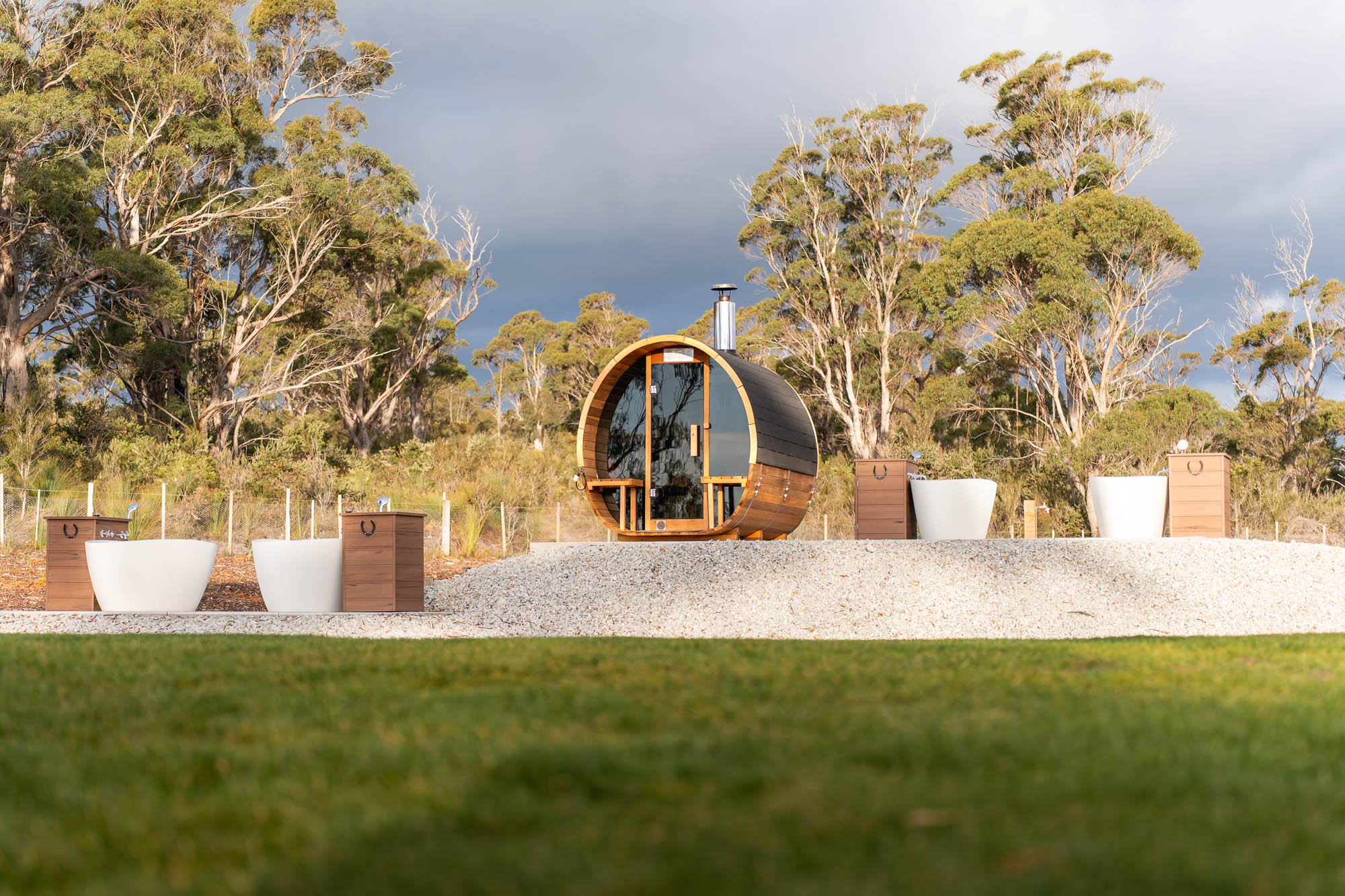 Outdoor wooden barrel sauna on a gravel platform surrounded by tall trees and garden tubs with wooden cabinets.