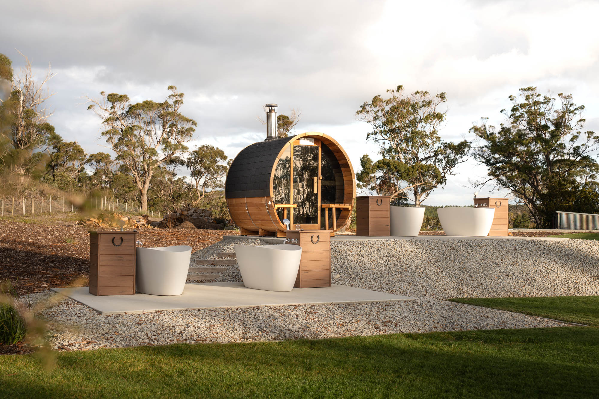 Outdoor spa area with a round wooden sauna cabin, two white soaking tubs, wooden faucets, and gravel landscaping surrounded by trees under a cloudy sky.