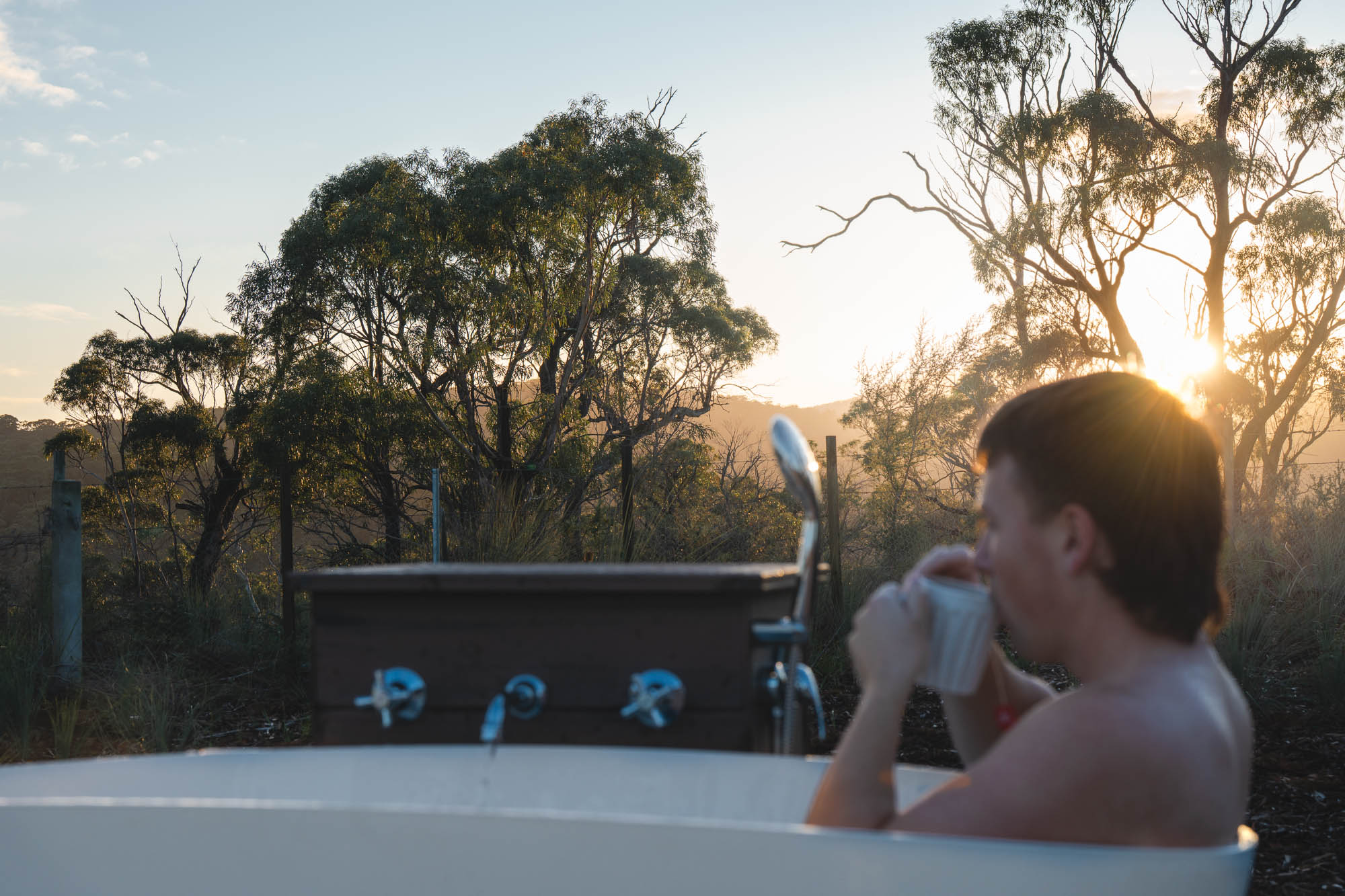 Person relaxing in an outdoor bathtub drinking from a mug at sunset with trees in the background.