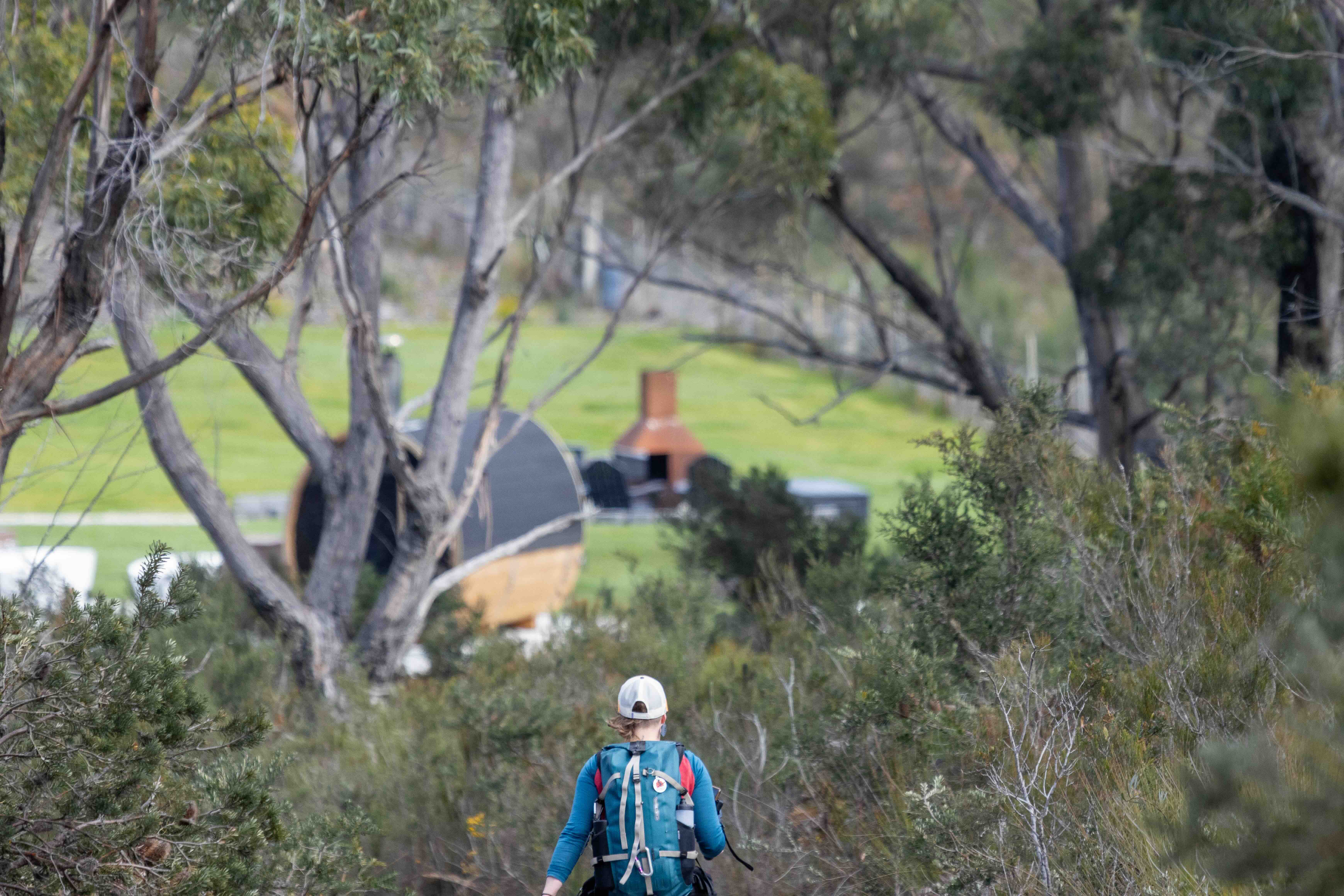 Person with a backpack walking on a trail surrounded by trees toward a grassy clearing with outdoor furniture and a brick barbecue.