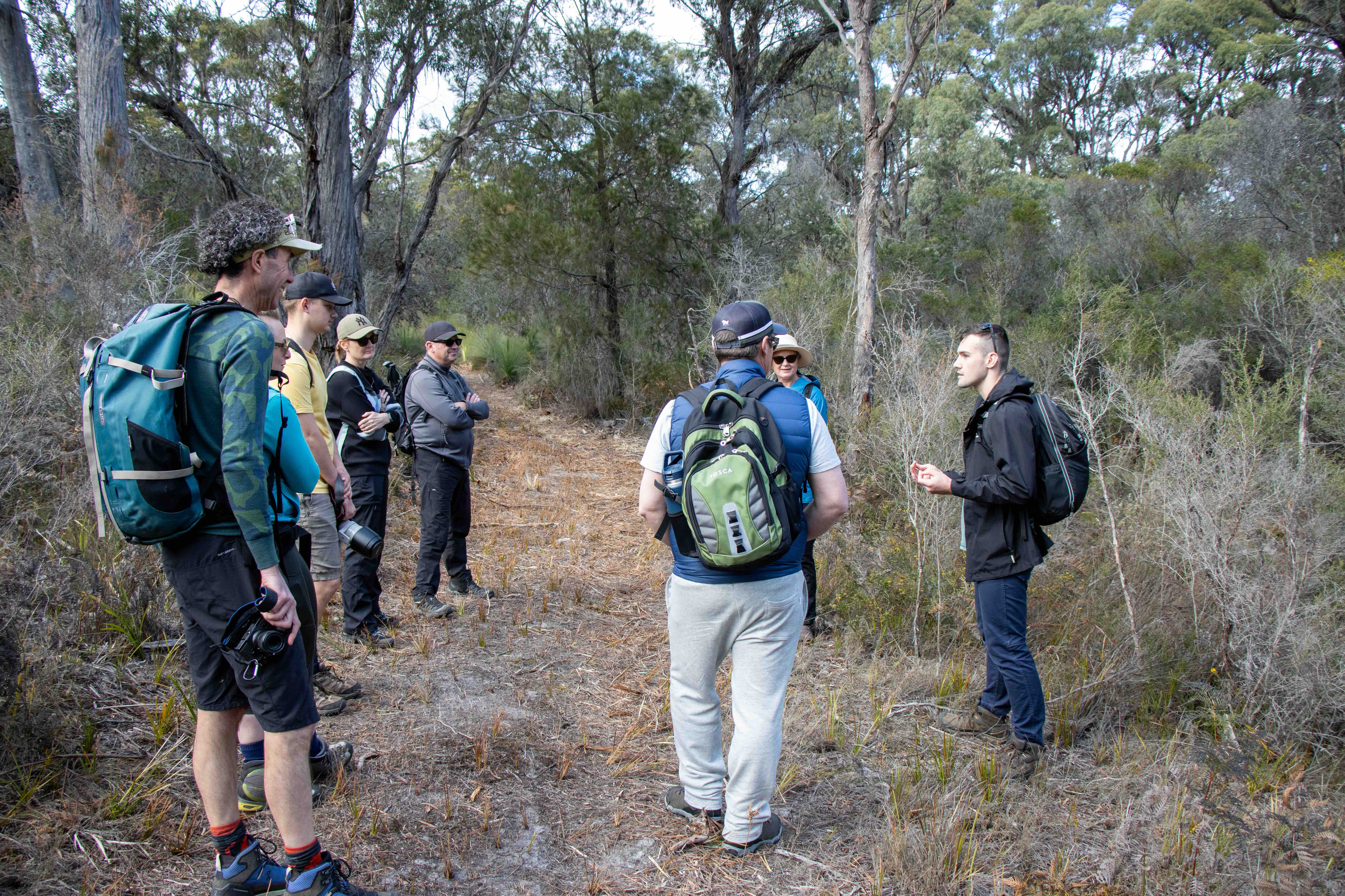 A group of eight people standing and listening to a guide speaking on a forest trail surrounded by tall trees and dry vegetation.