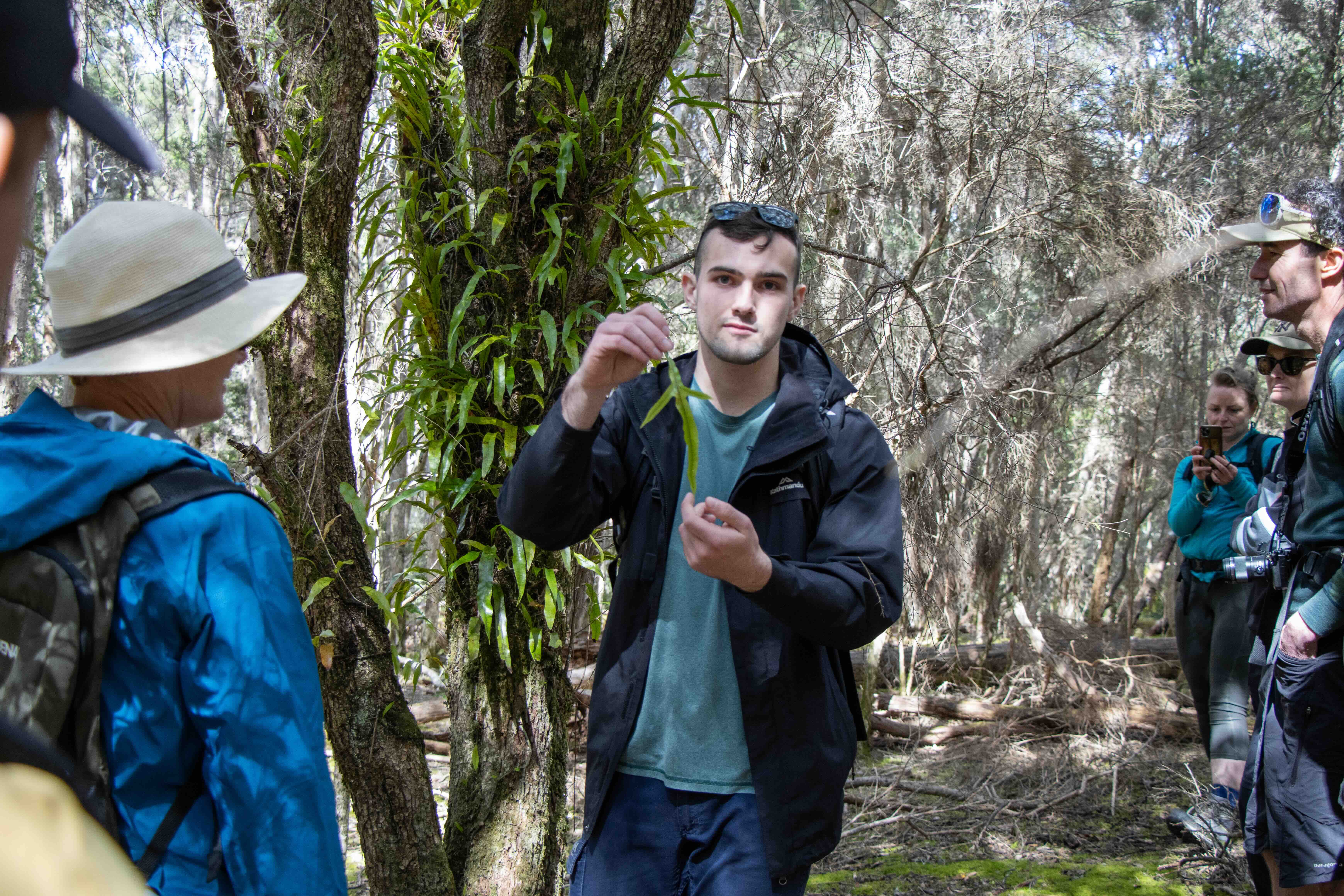 A man in a black jacket holding a fern leaf while a group listens attentively in a forest.
