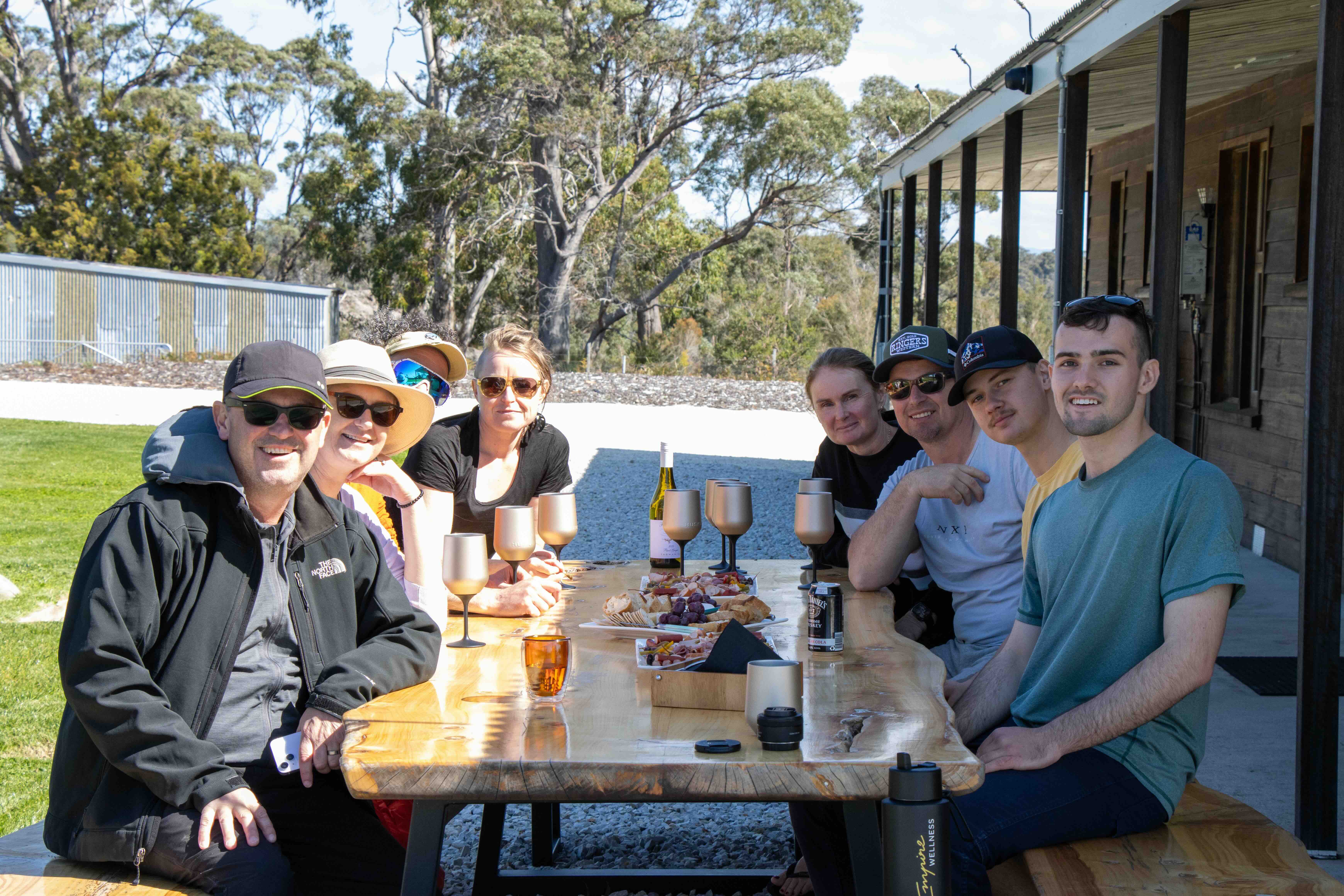 Group of seven people sitting outdoors around a wooden table with snacks and drinks, smiling at the camera.