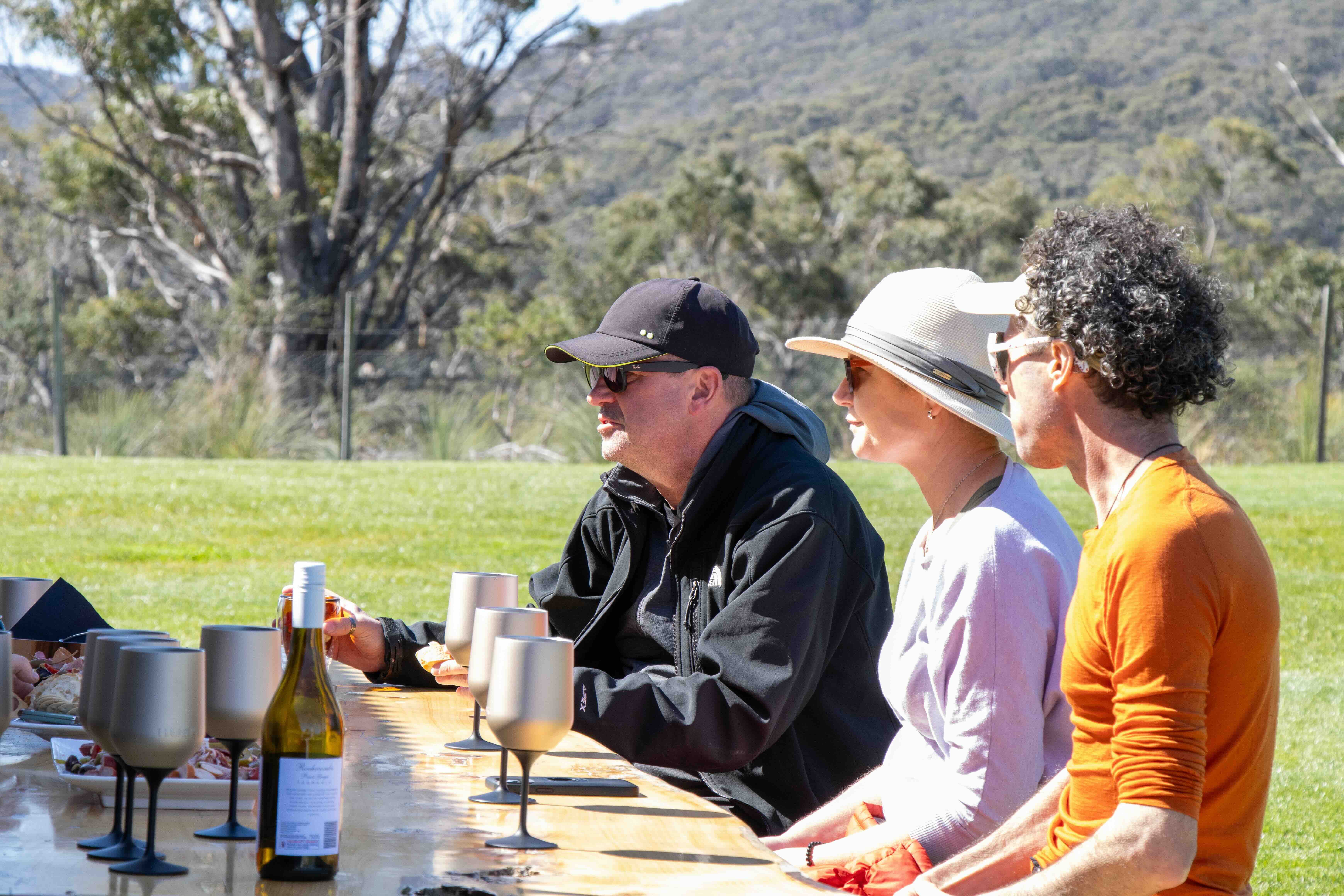 Three people sitting outdoors at a wooden table with grey wine glasses and a bottle, enjoying a sunny day in a grassy area with trees and hills in the background.