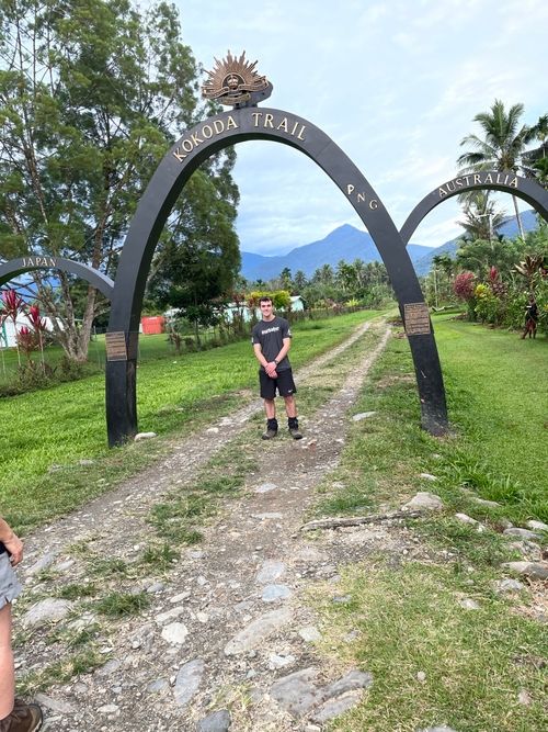 Person standing under a large archway marking the Kokoda Trail with mountains and lush greenery in the background.