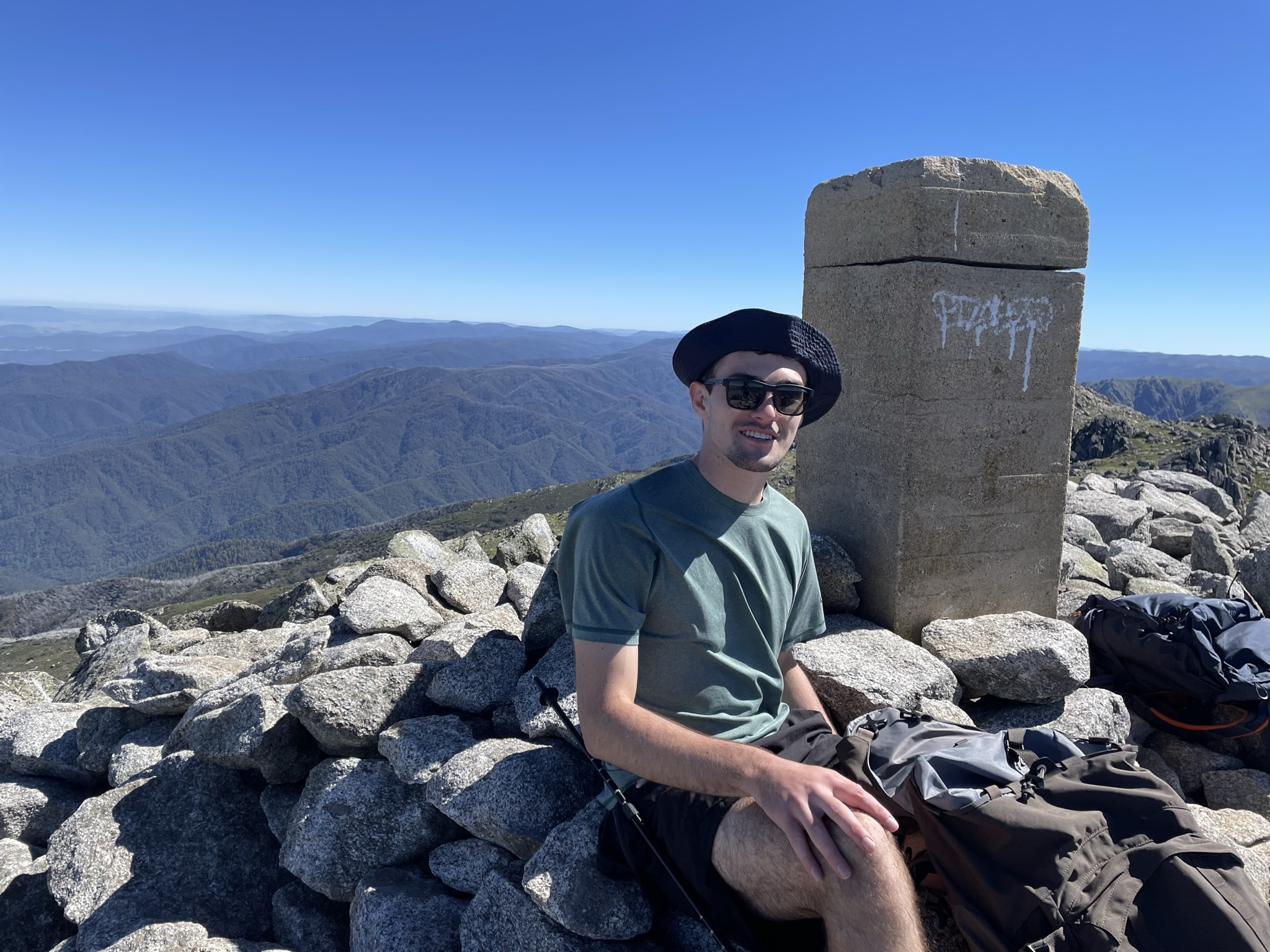 Man wearing sunglasses and a black beret sitting on rocky terrain near a stone pillar with mountain ranges in the background under a clear blue sky.